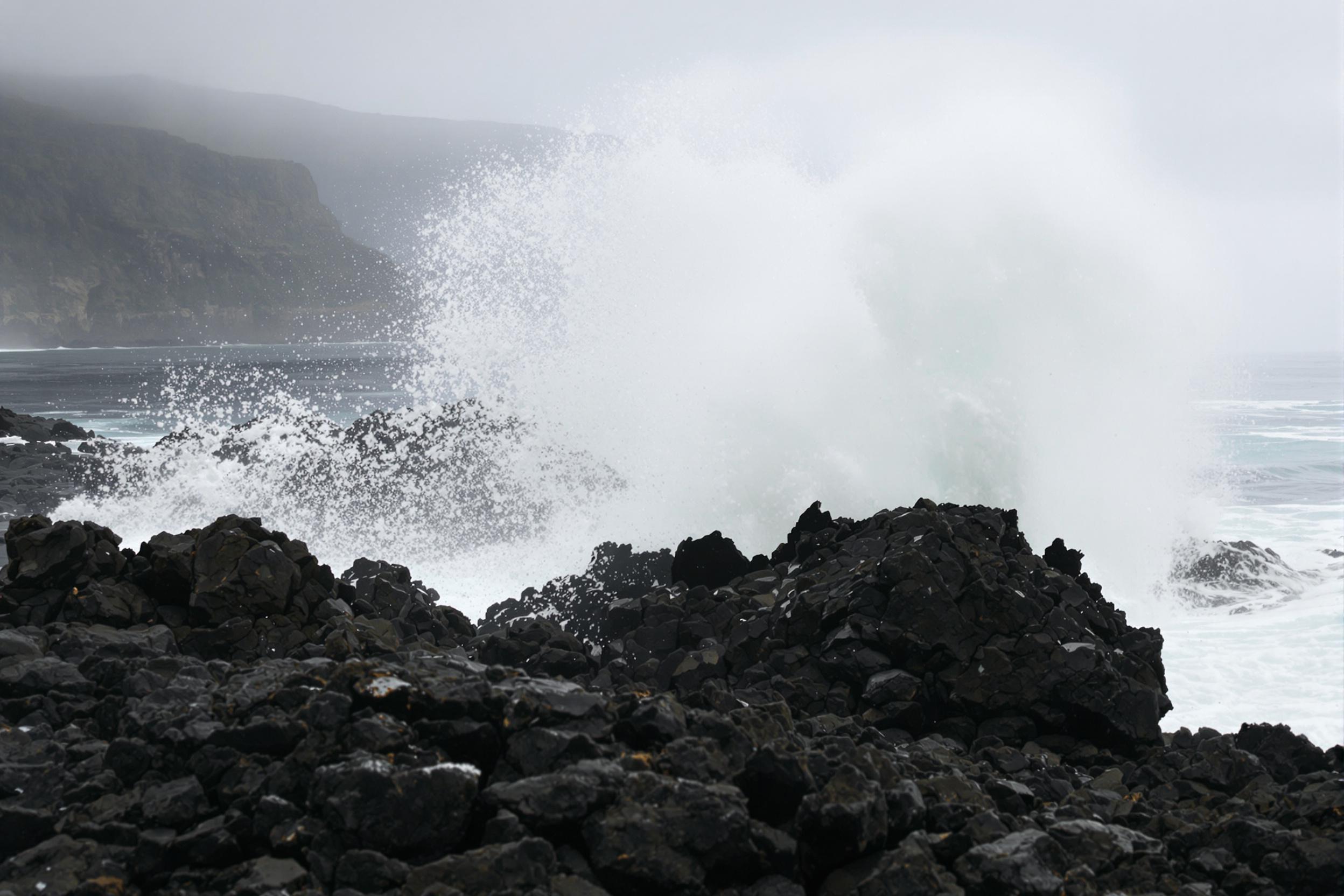 Forceful ocean waves crash violently against jagged black volcanic rocks, sending plumes of foamy white spray skyward. The dark, textured stones glisten under dim overcast skies, while blurred motion highlights the relentless energy of the surf. Mist softens the horizon, creating depth against the shadowy cliffs beyond.