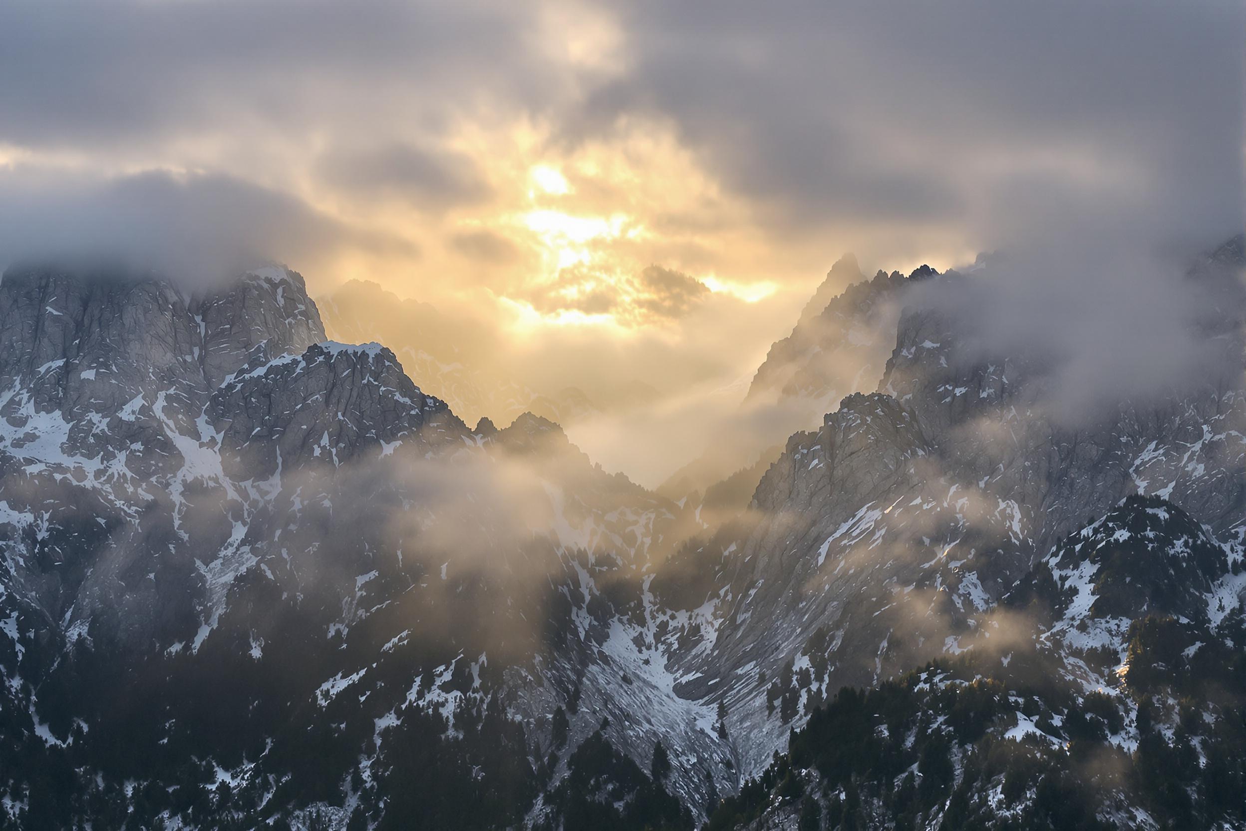 Capture the breathtaking beauty of a mountain range shrouded in morning mist, with golden sunlight piercing through the clouds. This panoramic shot showcases nature's grandeur, highlighting the interplay of light, shadow, and atmosphere across rugged peaks.