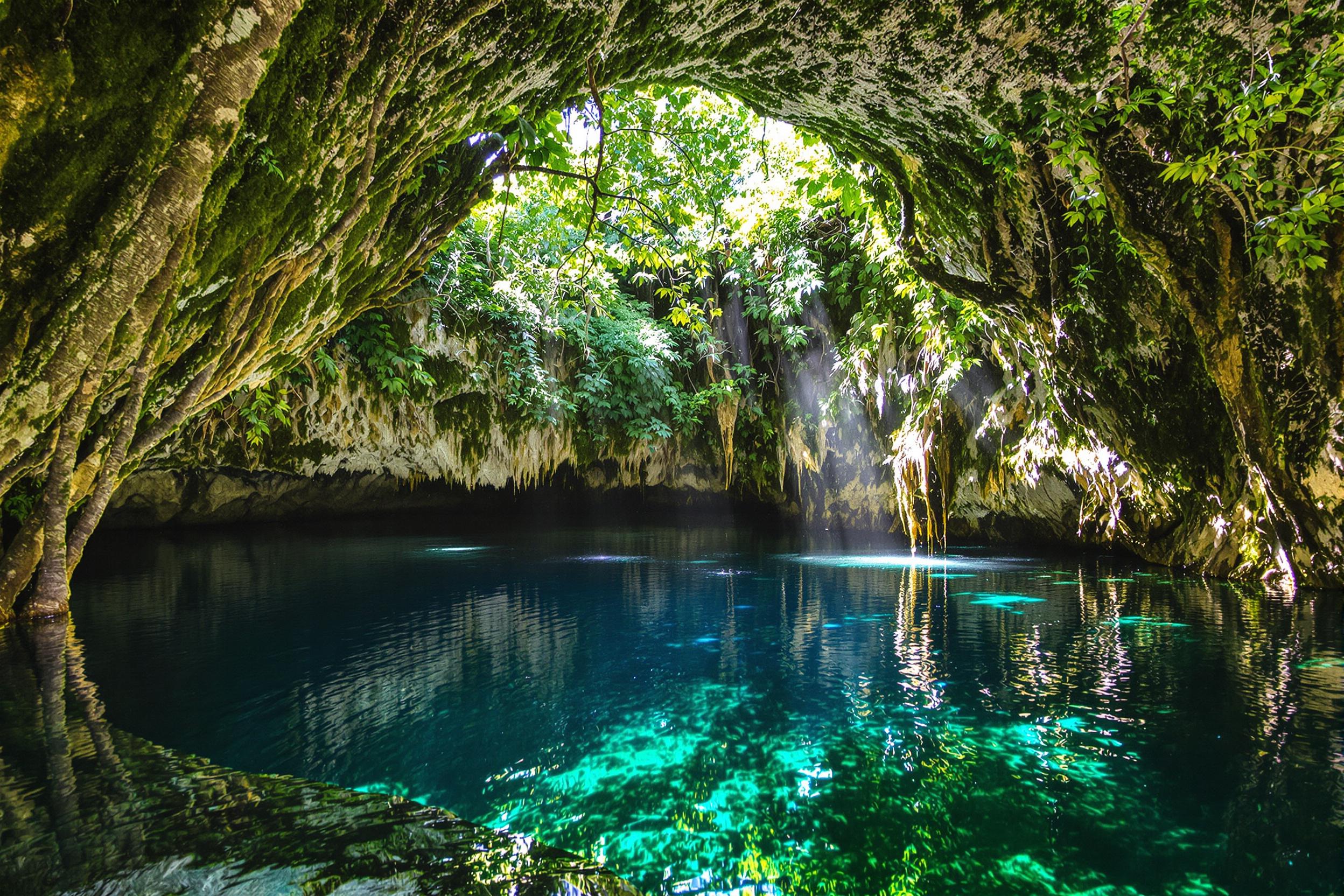 Discover a hidden gem in the Yucatan Peninsula: a mystical cenote surrounded by lush rainforest. Emerald waters reflect towering trees, while sunbeams pierce through the canopy, illuminating the moss-covered limestone walls. This serene oasis showcases nature's raw beauty and geological wonders.