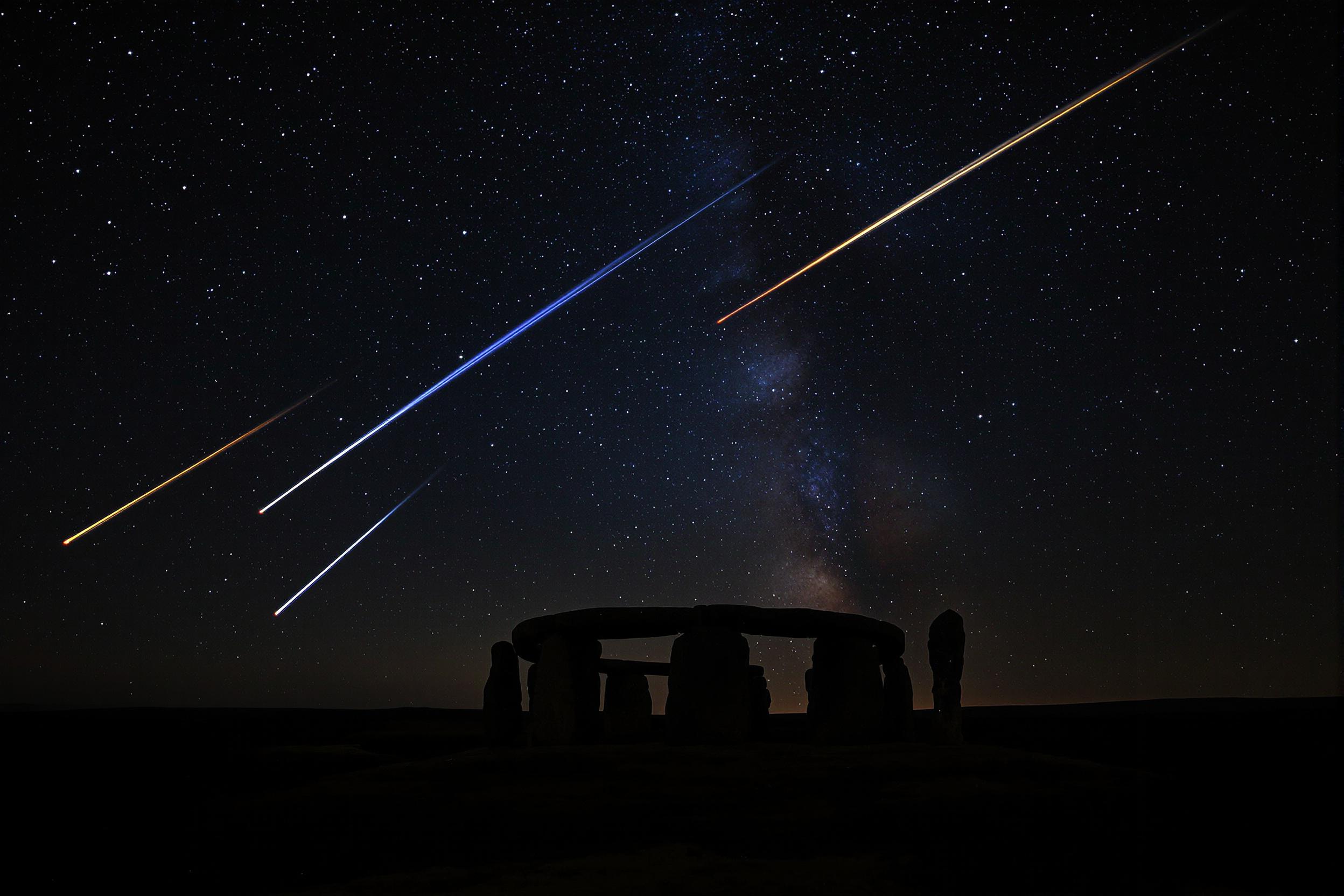 A mystical nighttime scene reveals an ancient stone circle silhouetted against a dark, clear sky. Radiant meteors streak across the Milky Way, adding drama to the expansive starscape. The arrangement offers sharp contrasts between glowing celestial phenomena and the shadowed terrain, bathed softly in faint starlight.