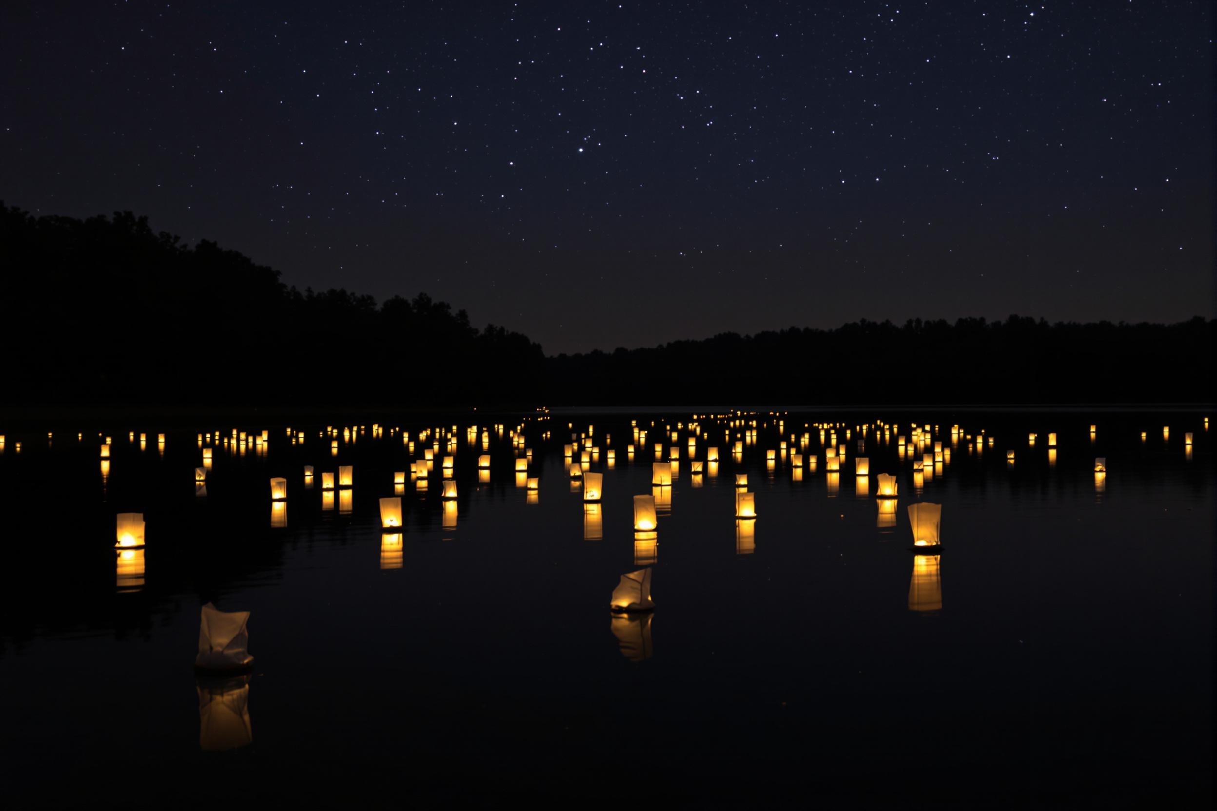 A serene scene unfolds at night as glowing lanterns float peacefully on a calm lake. The soft flickering light reflects on the glassy water, creating a magical ambiance. Silhouetted trees border the lake, their gentle outlines contrasting against the starry sky above. This tranquil environment invites contemplation and appreciation for nighttime beauty.