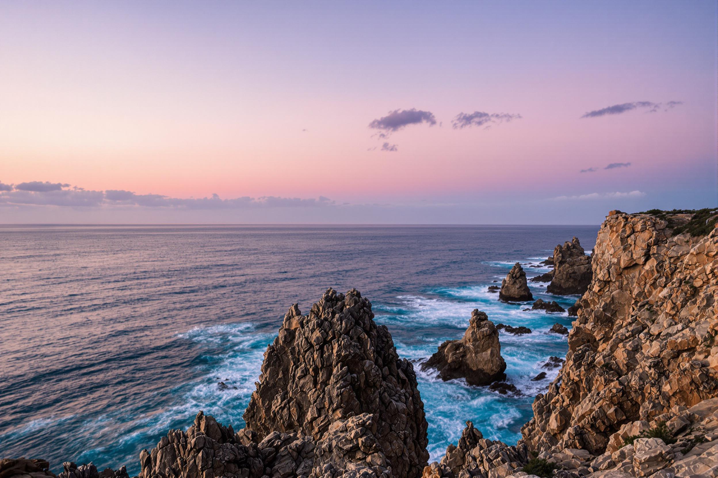 An expansive ocean view unravels from a dramatic rocky cliff at twilight. Jagged rocks protrude into the scene, contrasting against the vibrant blue of crashing waves below. The sky transitions from warm pink to deep indigo, reflecting off the water's surface. Wispy clouds scatter across the horizon, while gentle breezes ripple through the scene, enhancing its wild beauty.