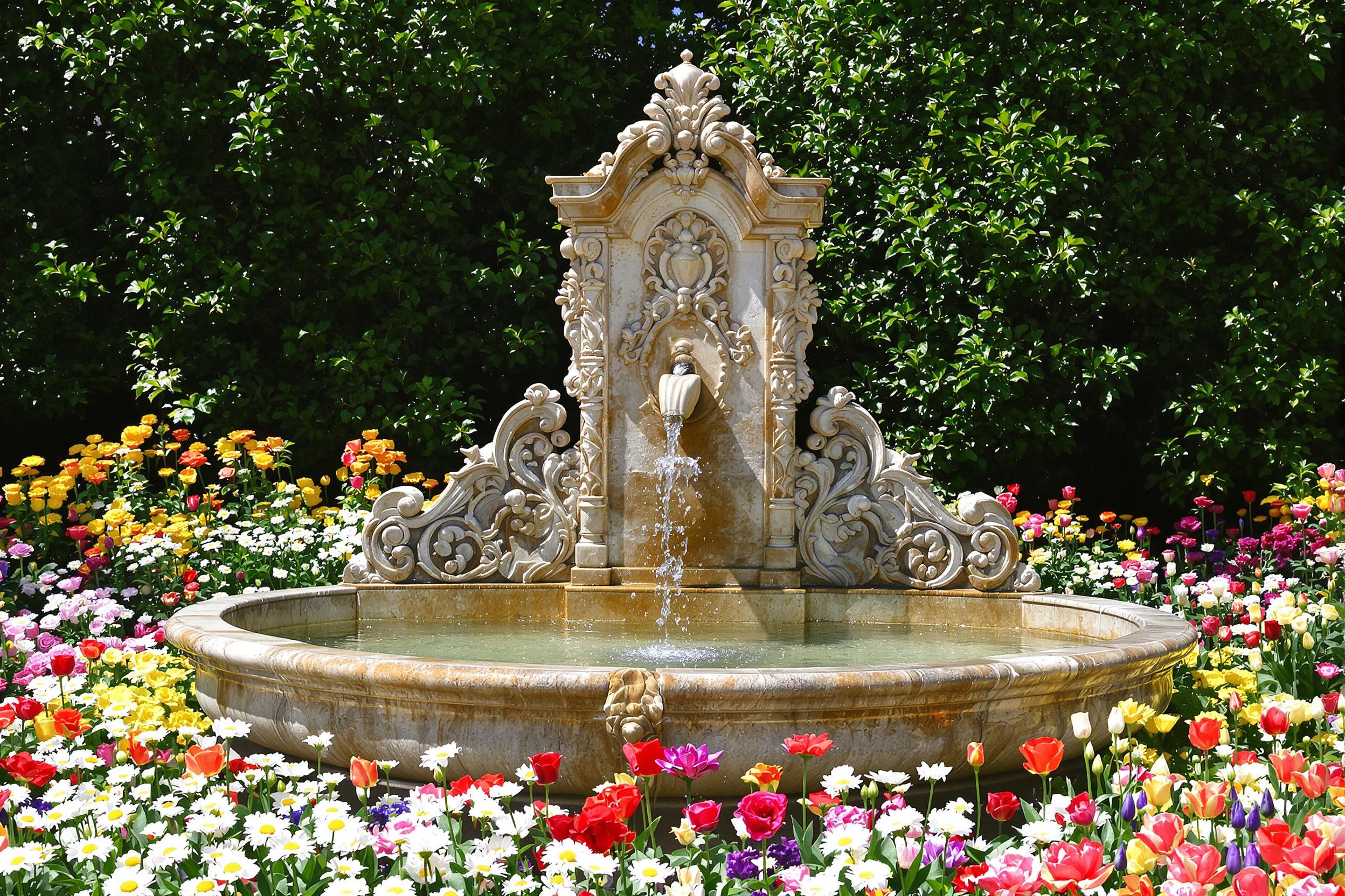 An ornate fountain stands elegantly at the center of a vibrant garden, surrounded by an array of blooming flowers. The fountain's intricately carved stone features flow water that glistens in the bright afternoon sun. Colorful petals of daisies, tulips, and roses frame the scene, while lush green foliage adds richness and depth.