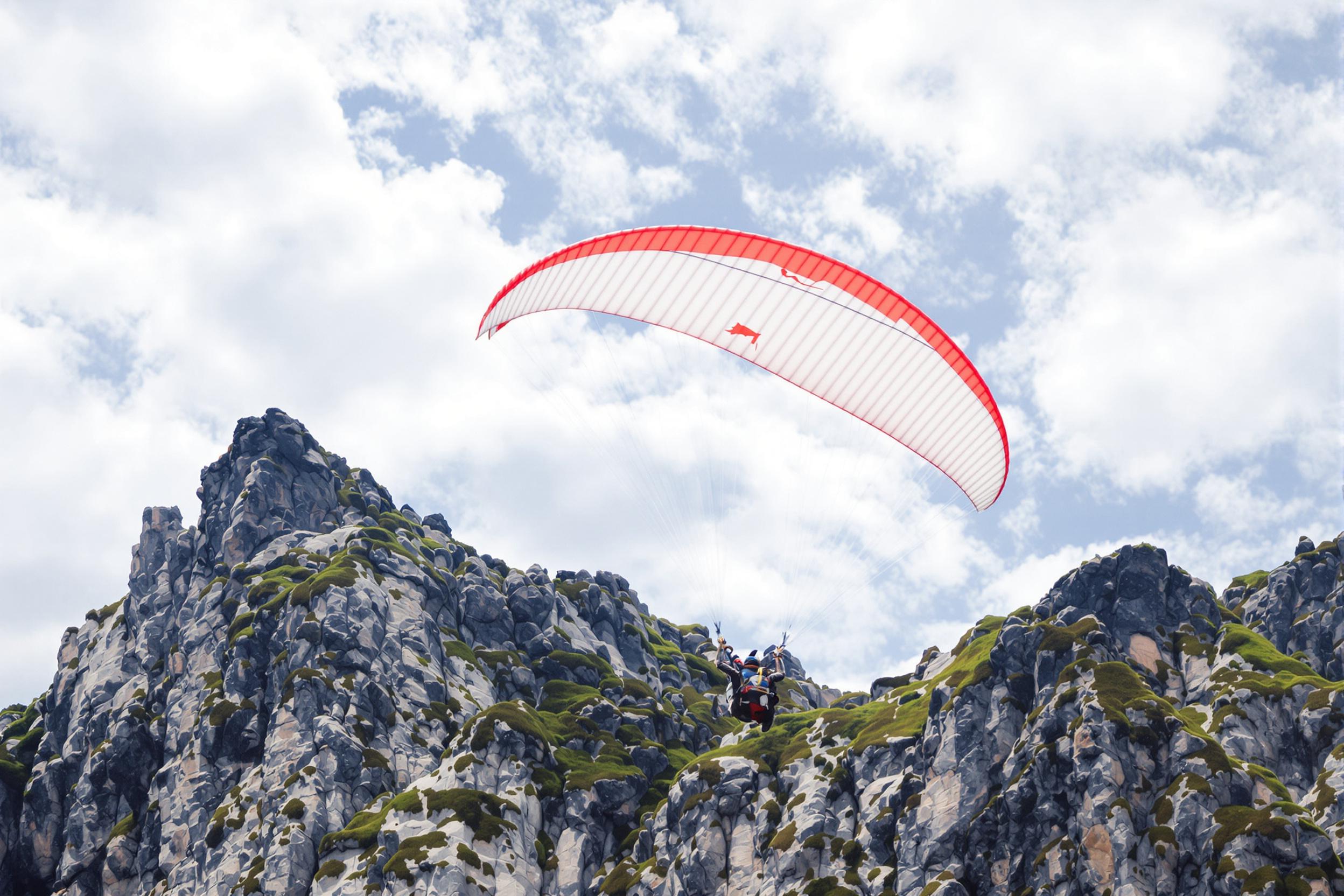 Against a backdrop of rugged, moss-covered mountains, a lone paraglider soars under a pale blue sky spotted with fluffy clouds. The vivid red-and-white parachute is taut in the breeze, contrasting sharply against the rocky cliffs below. Midday sunlight illuminates the scene, showcasing intricate folds in the fabric and jagged mountain textures.