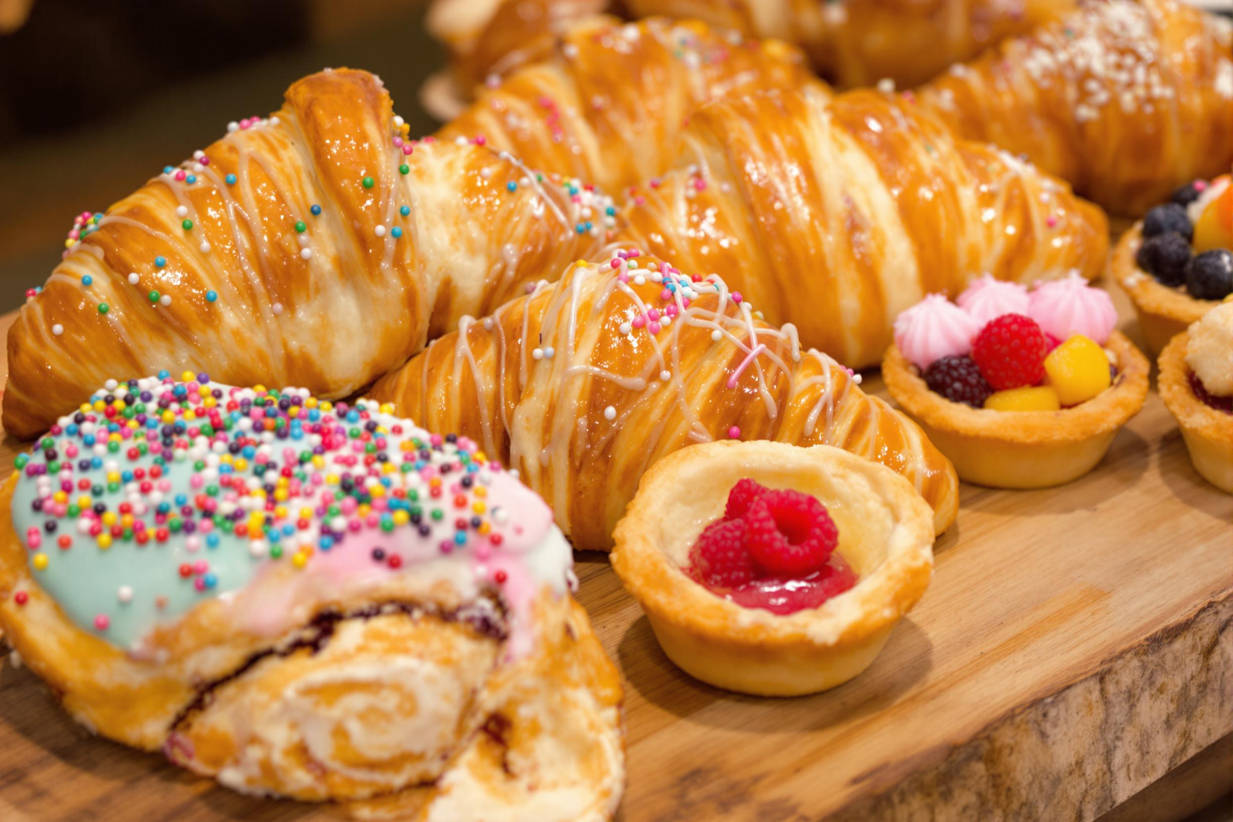 A close-up captures an array of golden-brown pastries artfully arranged on a rustic wooden board. Flaky croissants with glossy surfaces shine under soft morning light, while delicate éclairs boast colorful icing and intricate piping. Beside them, tartlets filled with bright fruits burst with color and texture, inviting indulgence. The warm hues evoke the cozy ambiance of a bustling patisserie.