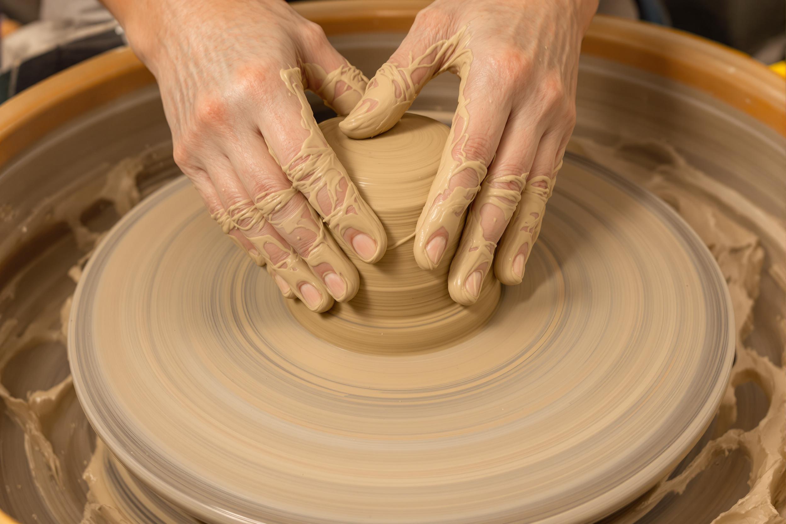 A tight frame captures hands elegantly molding a lump of wet clay onto a spinning pottery wheel. Streams of clay spin outward amidst splash marks as fingers press firmly to create defined ridges, revealing striking textures. Soft, warm lighting highlights earthy brown and beige hues, accentuating the scene's tactile richness.