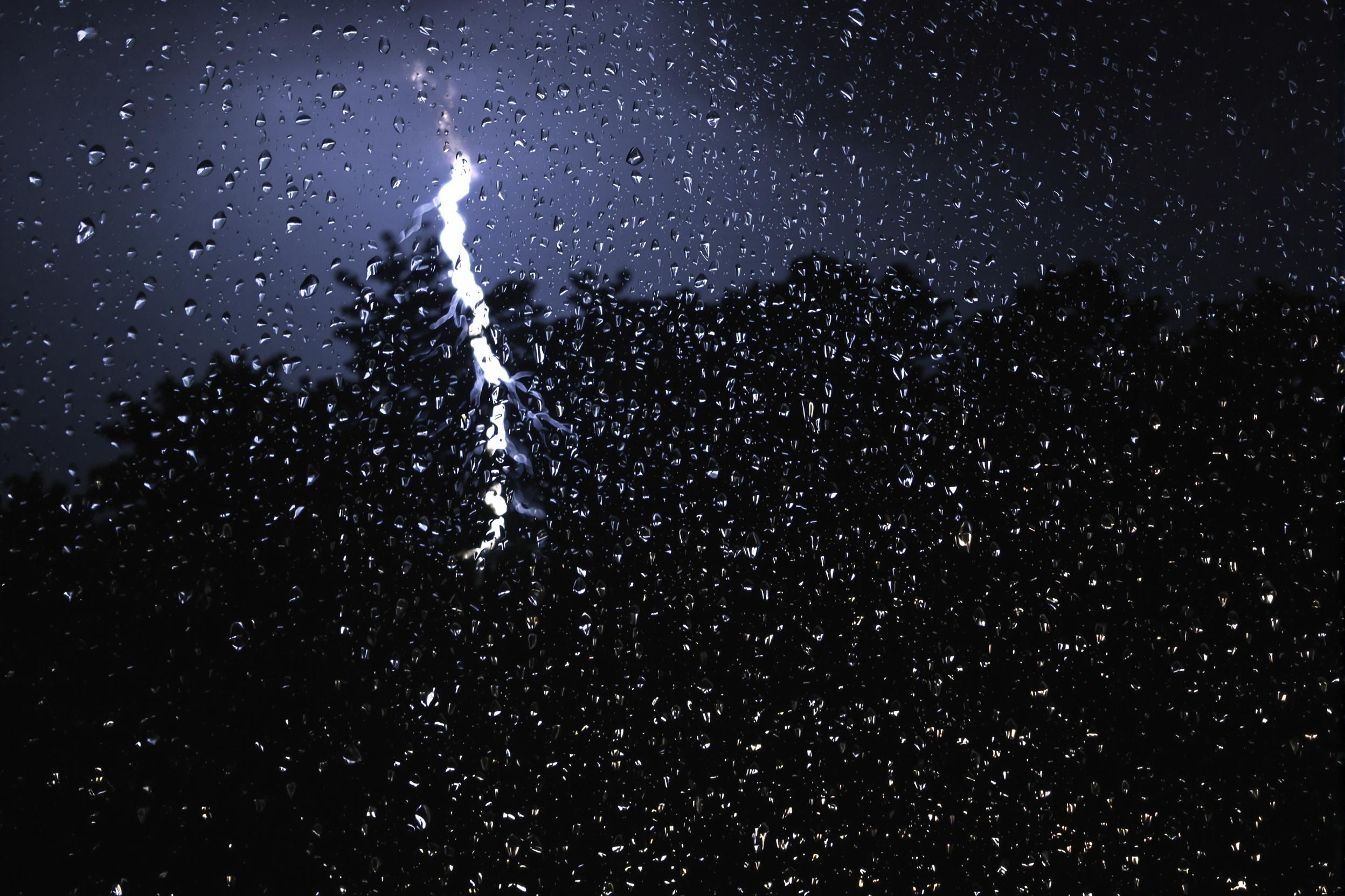 An extreme close-up of rain-soaked windowpanes during a thunderstorm. Glimmering droplets scatter as lightning momentarily illuminates the dim, distant storm scene. The rain streaks create a fluid, textured surface, contrasting with the softly blurred outlines of dark trees and turbulent skies beyond.