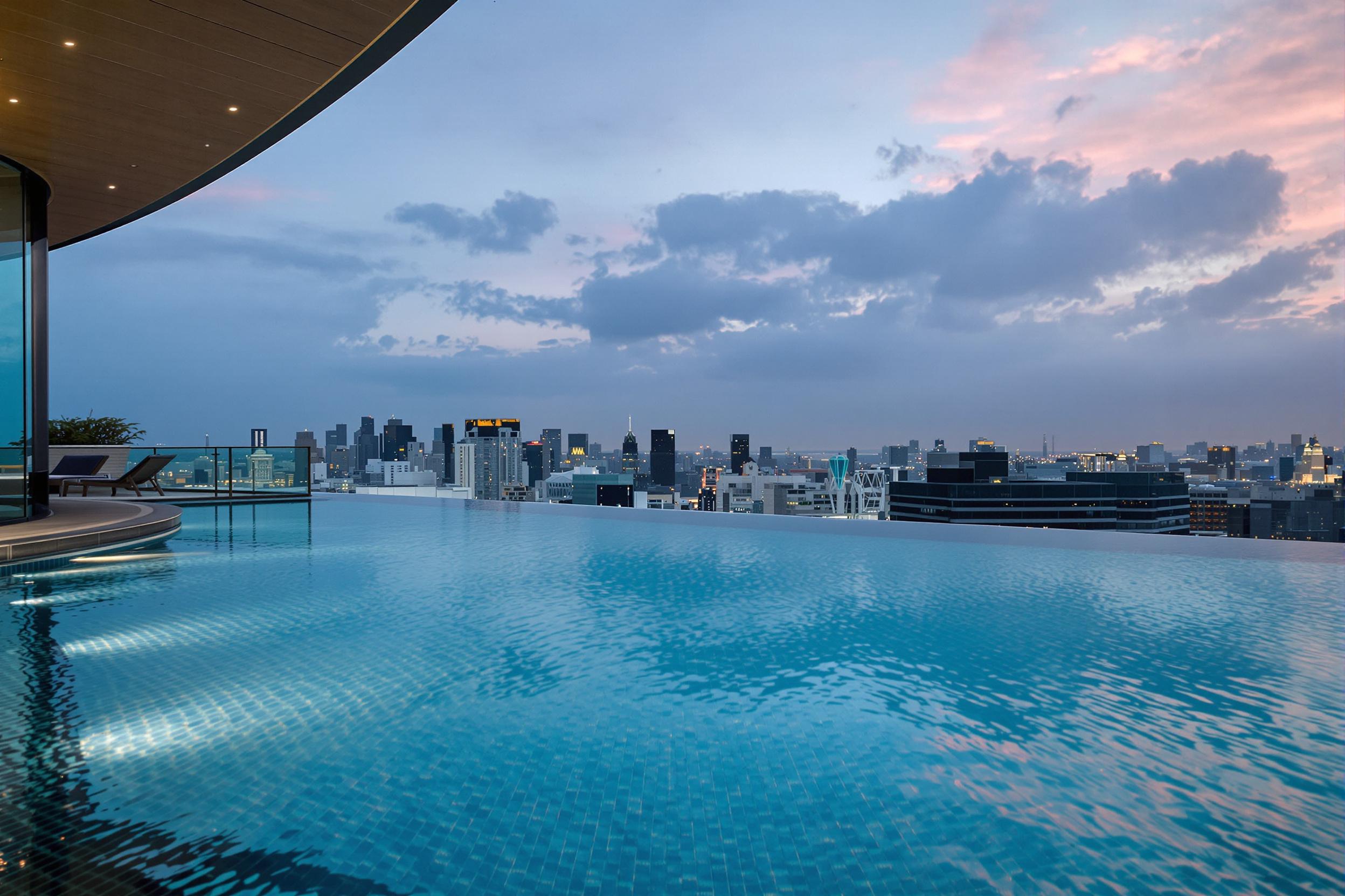 A serene rooftop infinity pool reflects pastel skies at dusk, framed by the modern geometry of high-rise buildings in a bustling city. Edges of the pool create the illusion of water blending into the distant skyline while subtle rippling patterns add texture to the glassy surface. Warm ambient light emphasizes geometric contrasts.