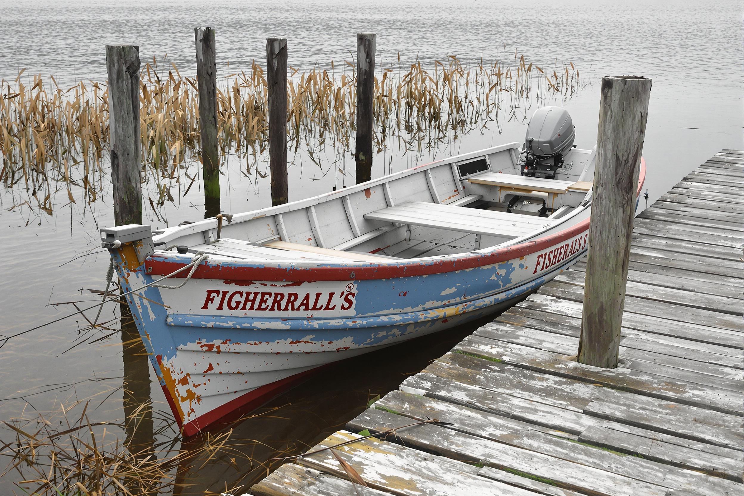 Moored by an old wooden pier, a weathered fisherman’s boat sits idle near a secluded lake. The textured wood planks display years of wear, complementing the chipped paint of the boat. Overcast light casts soft shadows on the calm, reflective water, while reeds along the shore add subtle vertical accents to the tranquil composition.