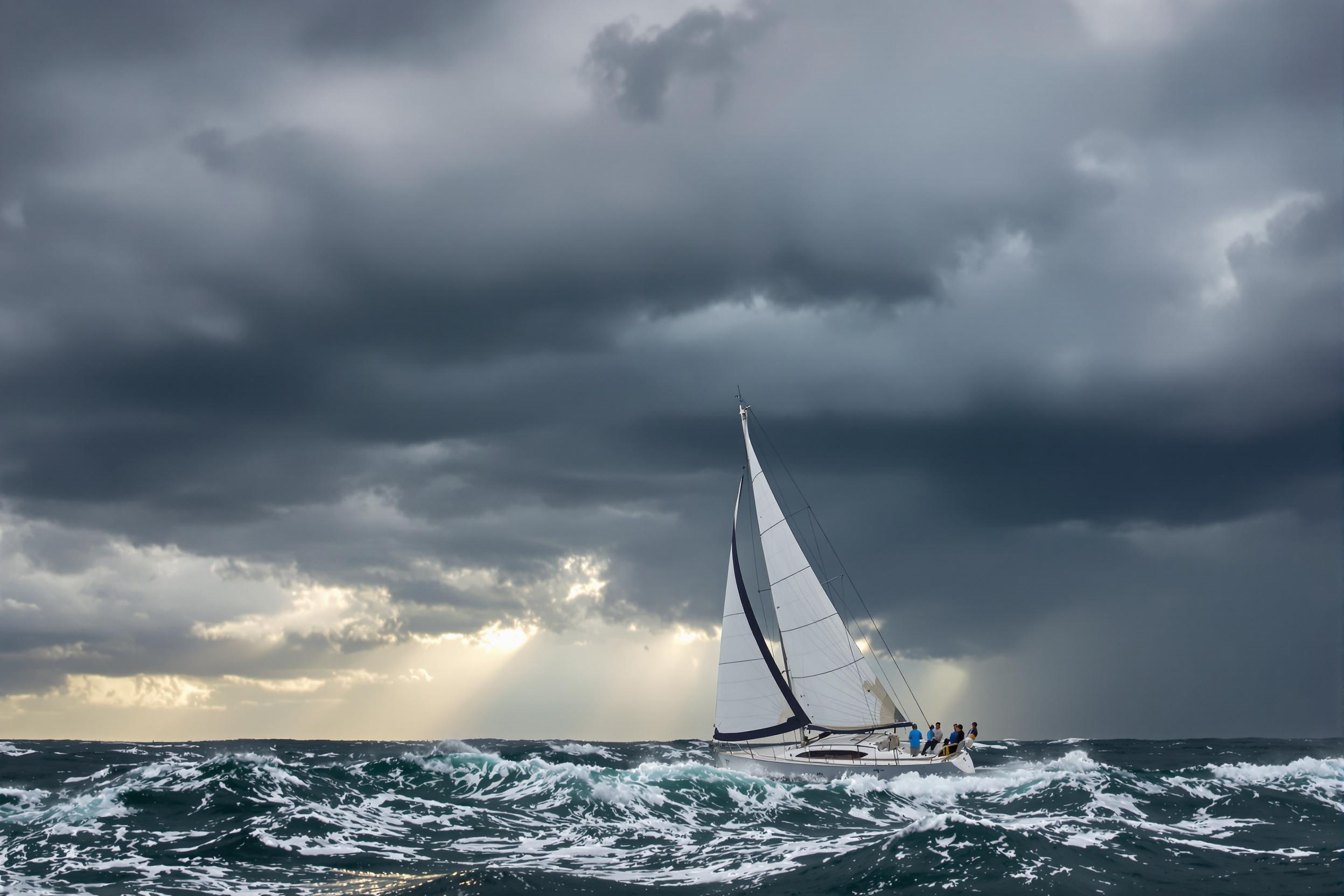 A sailboat slices through turbulent waves under ominous dark clouds. Gusts fill its billowing white sails as glimmers of sunlight pierce the brooding sky. The wide frame captures the dynamic interplay between sharp rippling water textures and expressive stormy elements overhead.