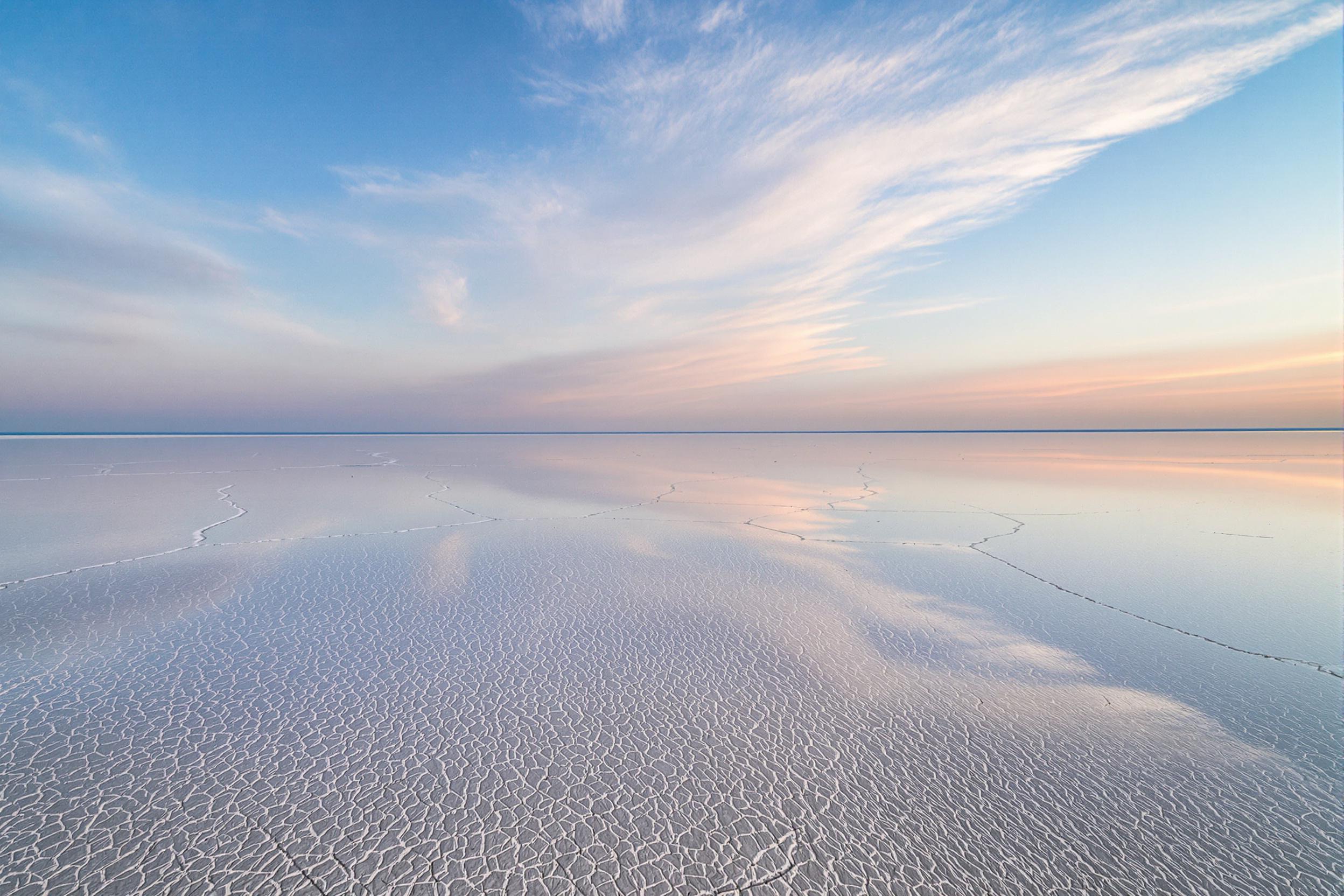 A vast expanse of salt flats stretches into the horizon, their crystalline surface creating mirror-like reflections underneath soft dawn light. Patterns of cracked earth break up sections of smooth glassy areas, where ripples from distant wind disrupt the otherwise perfect reflections of a pale blue and pink sky above.