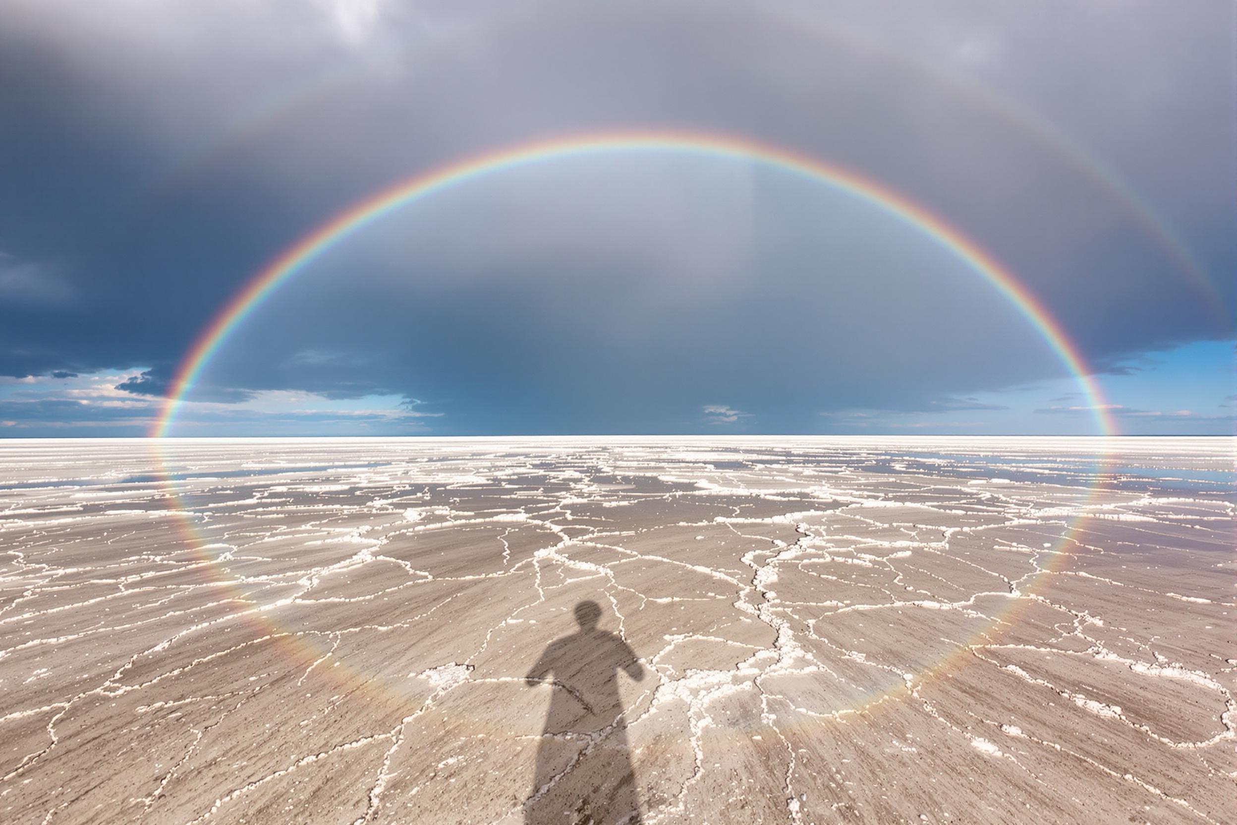 An expansive salt flat stretches under a vibrant double rainbow arching across the sky. The wide-angle composition captures gleaming water puddles reflecting both rainbows, intensifying a spectrum of colors against the muted earthy tones of the flat. Dramatic storm clouds linger, adding depth and contrast to the ethereal scene.