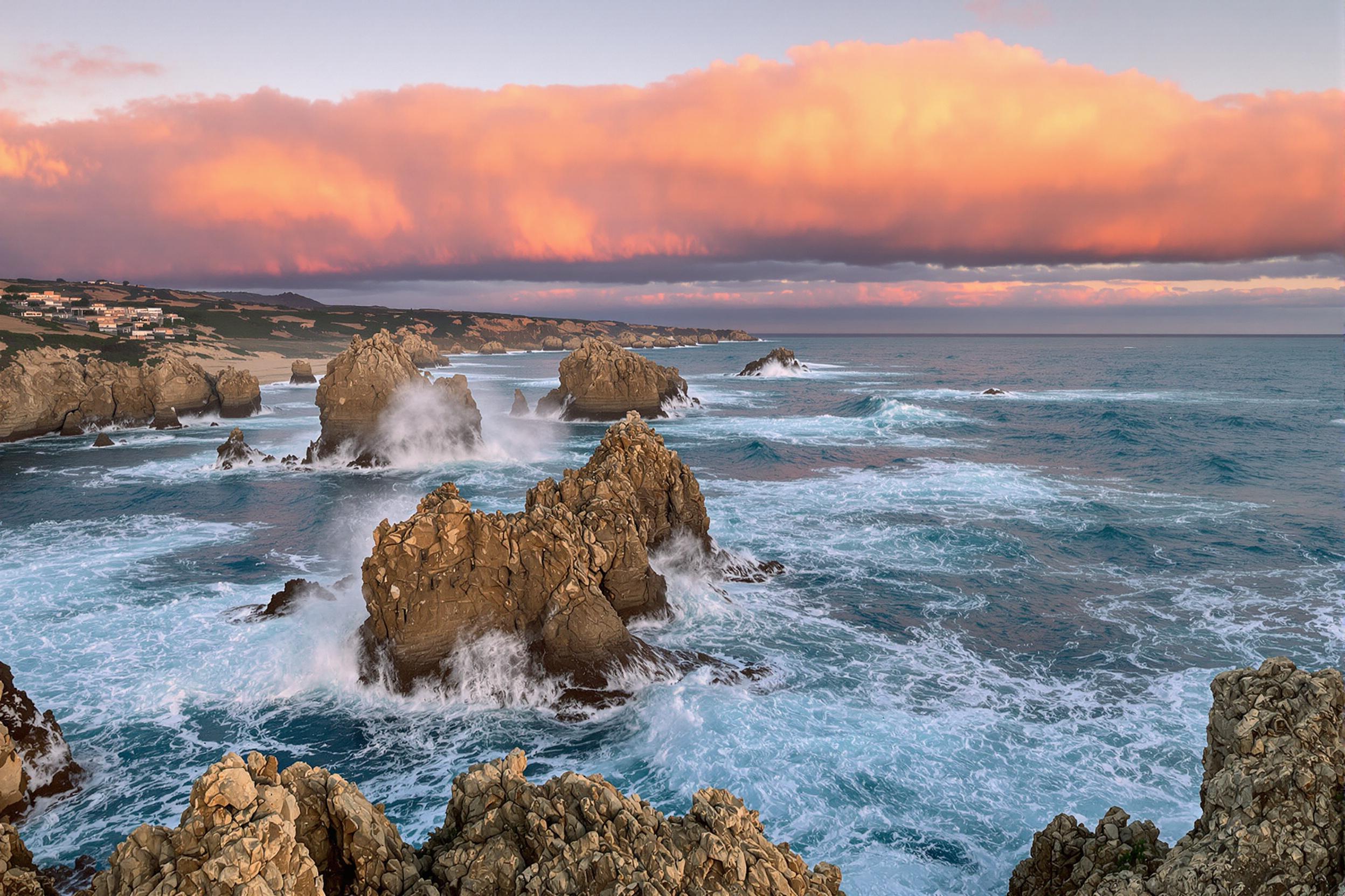 A breathtaking seascape captures the power of nature as waves crash against jagged cliffs at sunset. The dynamic movement of frothy white water contrasts with deep blue ocean tones. Sunlight washes over the rugged landscape, casting warm golden hues that illuminate the rocks. Layers of clouds reflect pink and orange shades above, creating a dramatic backdrop.