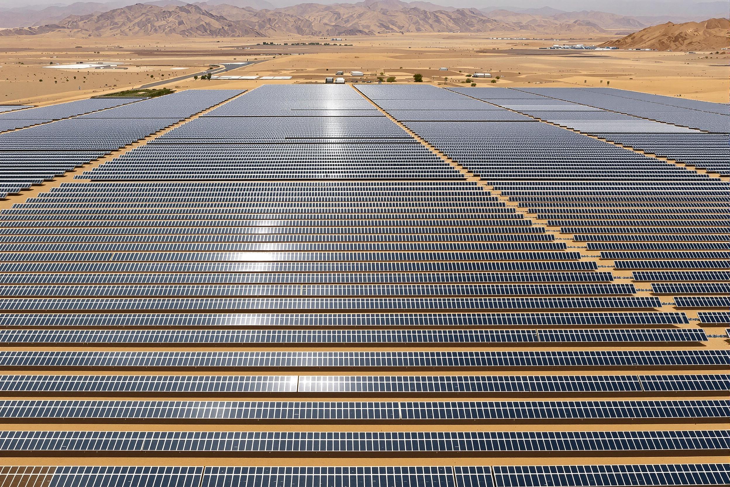 Arrays of gleaming solar panels stretch endlessly over arid desert terrain, perfectly aligned in crisp geometric rows. Captured from above, their reflective surfaces glisten under bright midday sunlight, casting distinct shadows on the golden-hued sand below. Vast ridges frame the distant horizon, completing this grand renewable energy landscape.