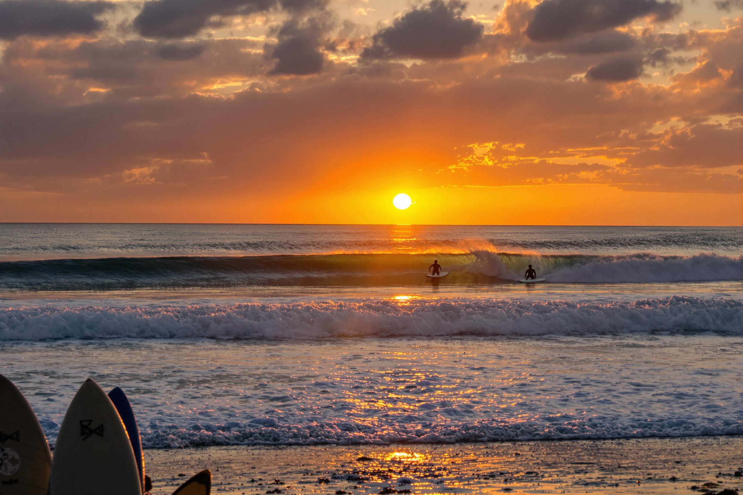 Surfers carve through rolling waves under a stunning sunset backdrop. The golden and orange hues reflect off the water, creating a vibrant glow. In the foreground, surfboards are positioned in the sand, giving context to the energetic scene. Framed by gentle waves and the setting sun, this idyllic beach captures the essence of outdoor adventure.