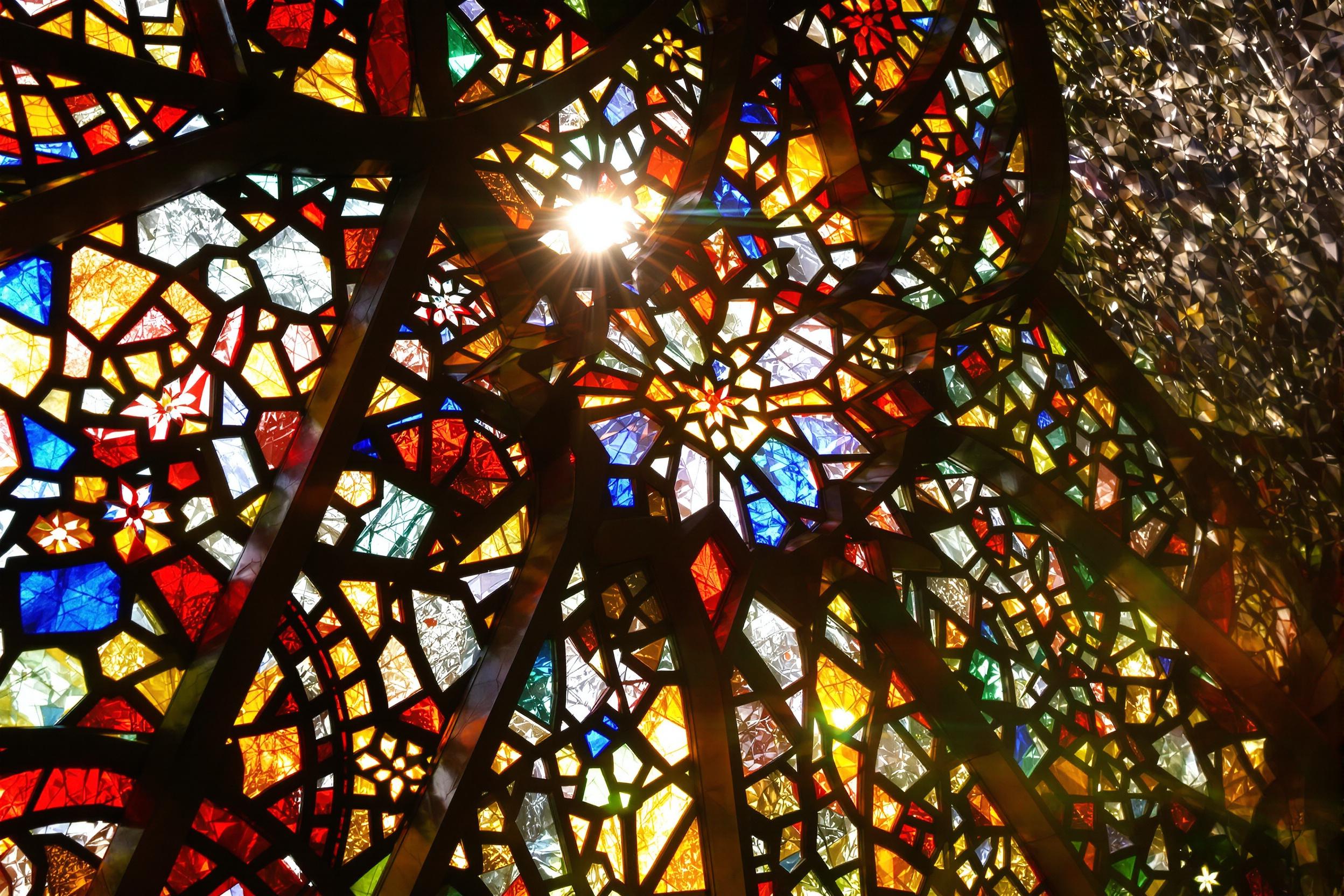 An extreme close-up highlights the intricate beauty of stained glass. Sunlight streaming through vividly illuminates reds, blues, yellows, and greens bordered by dark metallic latticework. Curved floral and geometric motifs interlock seamlessly, casting dappled multicolored reflections onto surrounding stone. Every fragment glimmers in sharp detail.