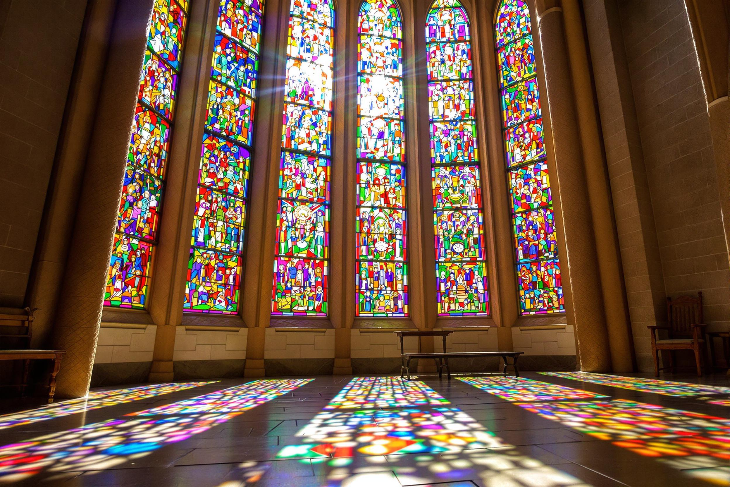 An exquisite stained glass window fills the wall of a gothic church, showcasing a vivid array of colors formed into intricate patterns. Bright afternoon sunlight streams through, casting colorful reflections onto the stone floor. The detailed craftsmanship emphasizes the beauty and artistry, while the soft shadows add depth to this captivating scene.