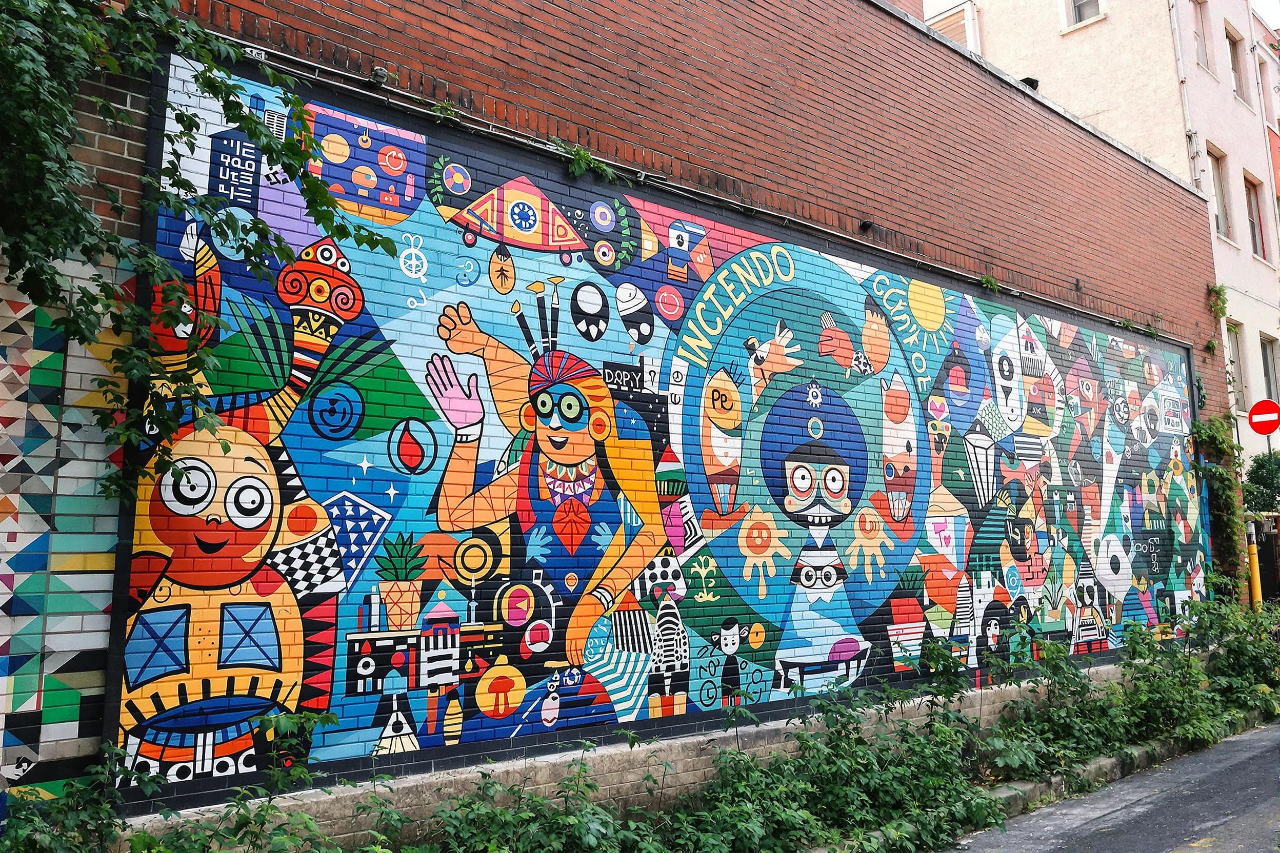 A colorful street mural brings an urban wall to life, depicting intricate designs that celebrate cultural heritage. Bright blues, oranges, and greens dance across the canvas, showcasing artistic characters and patterns. Set against a muted brick background, the mural's vibrancy is enhanced by surrounding greenery and urban elements that invite exploration of creativity in public spaces.