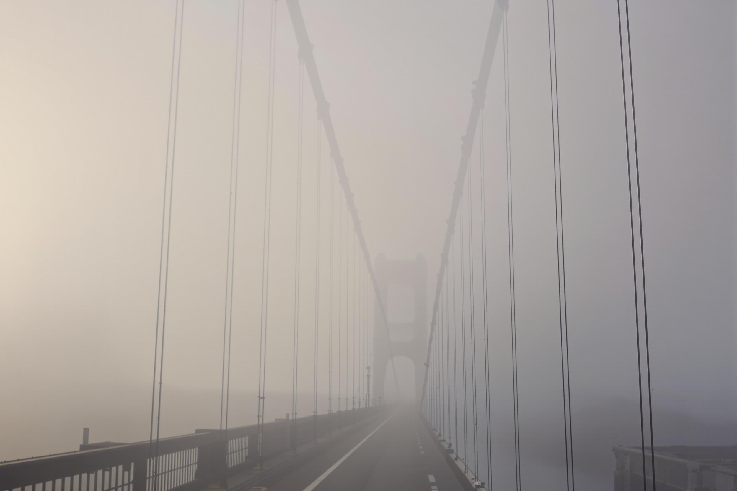 An iconic suspension bridge fades gradually into dense fog, captured from a wide, low perspective amid muted early-morning light. Steel cables form leading lines that vanish into the mist, creating a sense of vastness and mystery. Subtle texture and desaturated tones enhance the cinematic quality, evoking an ethereal urban aesthetic.