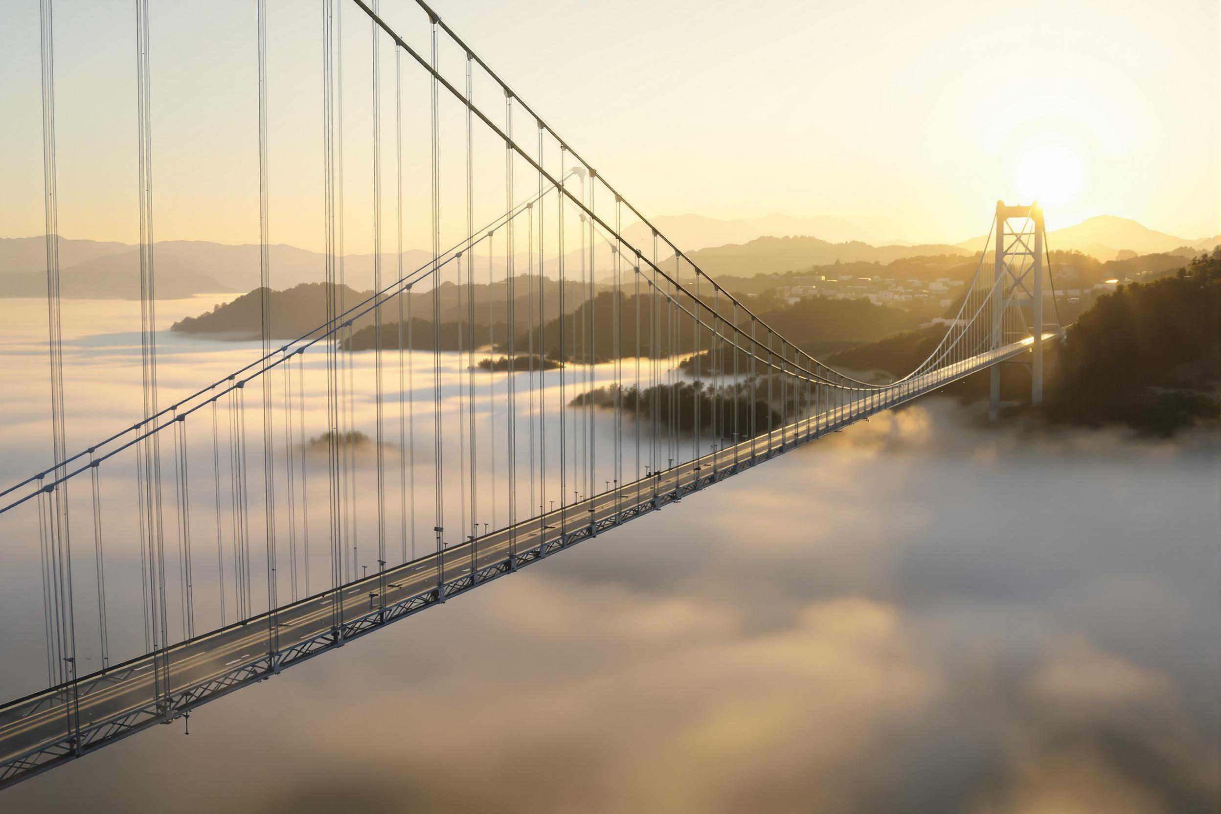 Vast suspension bridge arches majestically over a fog-filled valley at sunrise. Golden sunlight softly illuminates the bridge’s towering structure, creating stark contrast against the dense mist obscuring the river below. Thin cables fan out symmetrically, drawing the eye toward the horizon where hazy mountains rise faintly under warming hues of dawn.