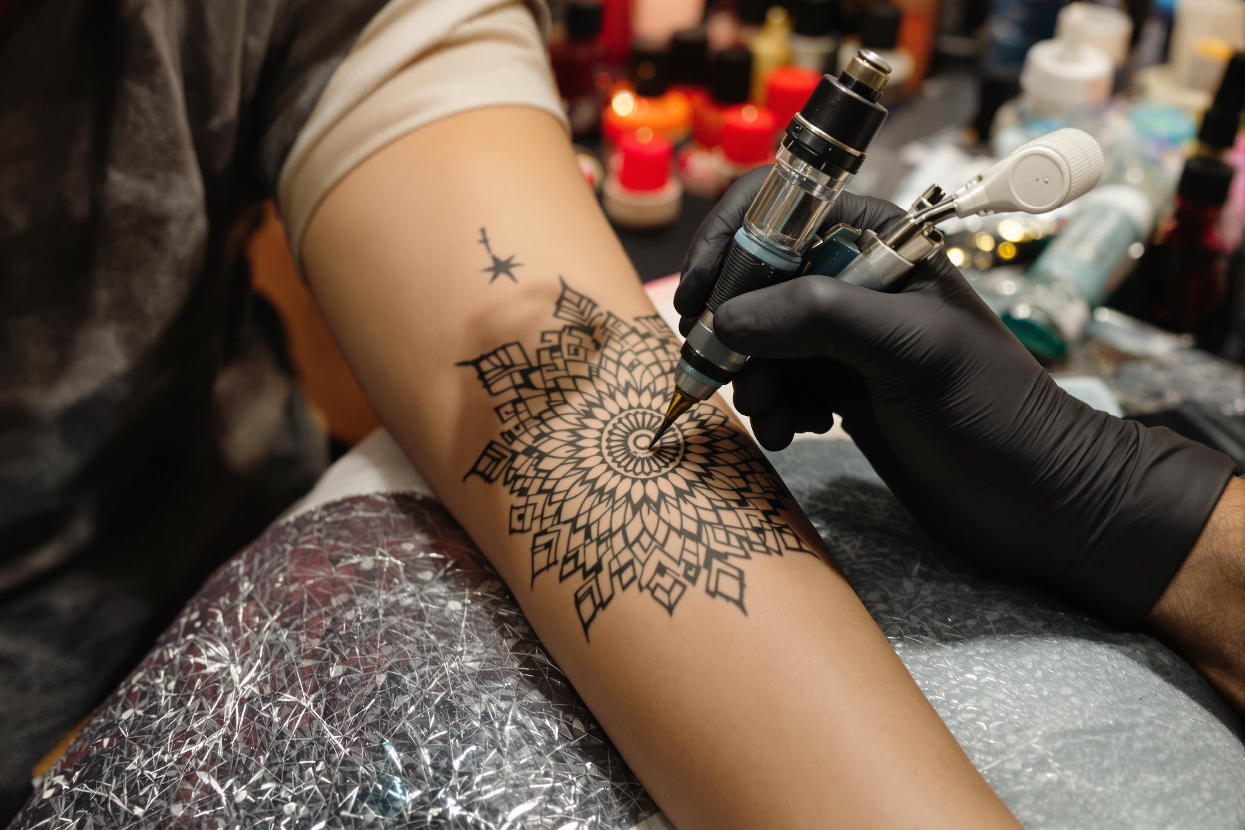 A close-up shot captures the intricate process of a mandala tattoo being applied on a client's arm. The artist’s skilled hands hold the tattoo machine with precision, creating delicate patterns on the skin. Soft, warm light filters in, illuminating the shades of ink while various tattoo supplies are arranged within reach, enhancing the creative atmosphere.