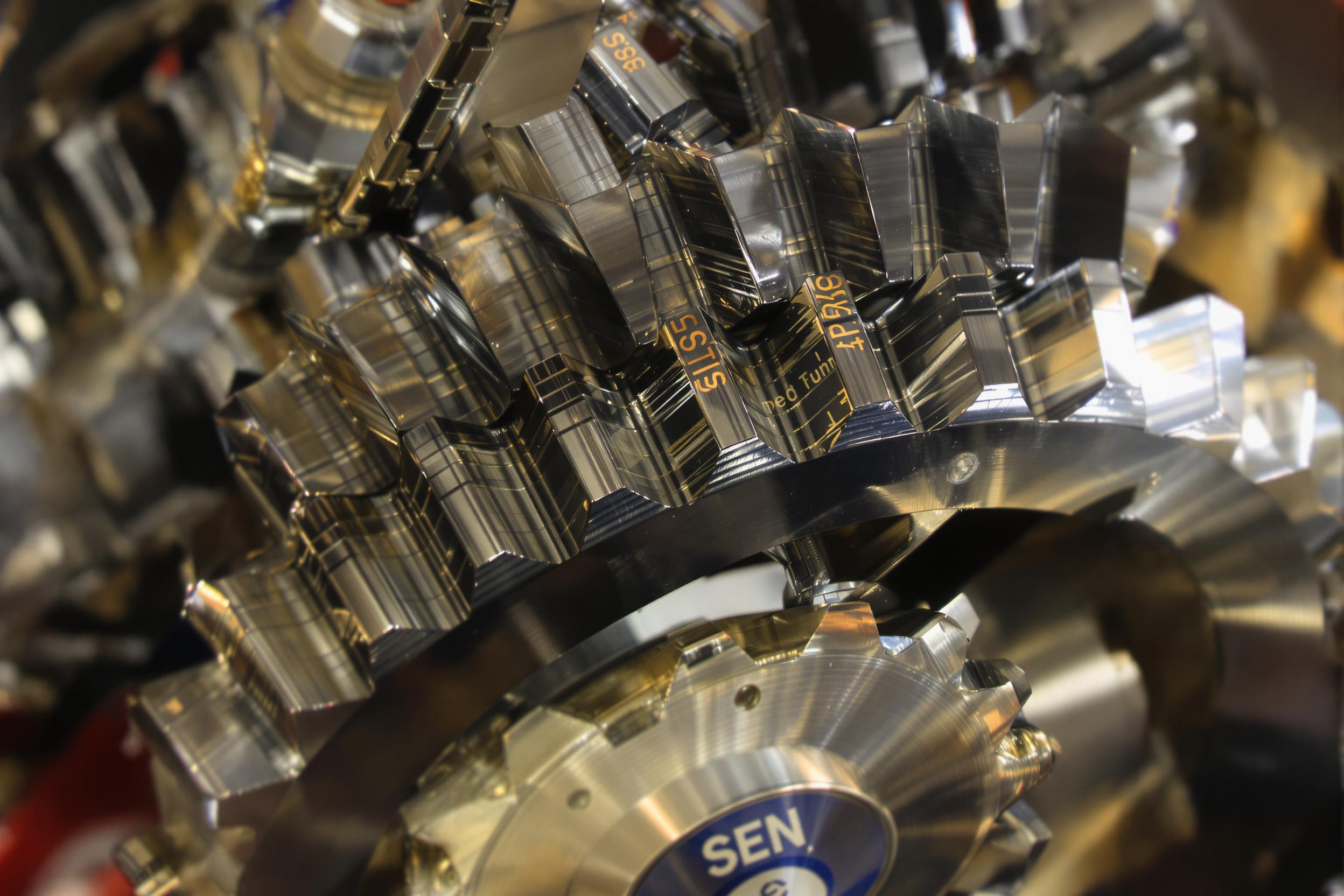 Close-up photograph of interlocking metal gears, showcasing the intricate patterns and textures of industrial machinery. Polished steel surfaces reflect ambient light, emphasizing the precise engineering and mechanical beauty.