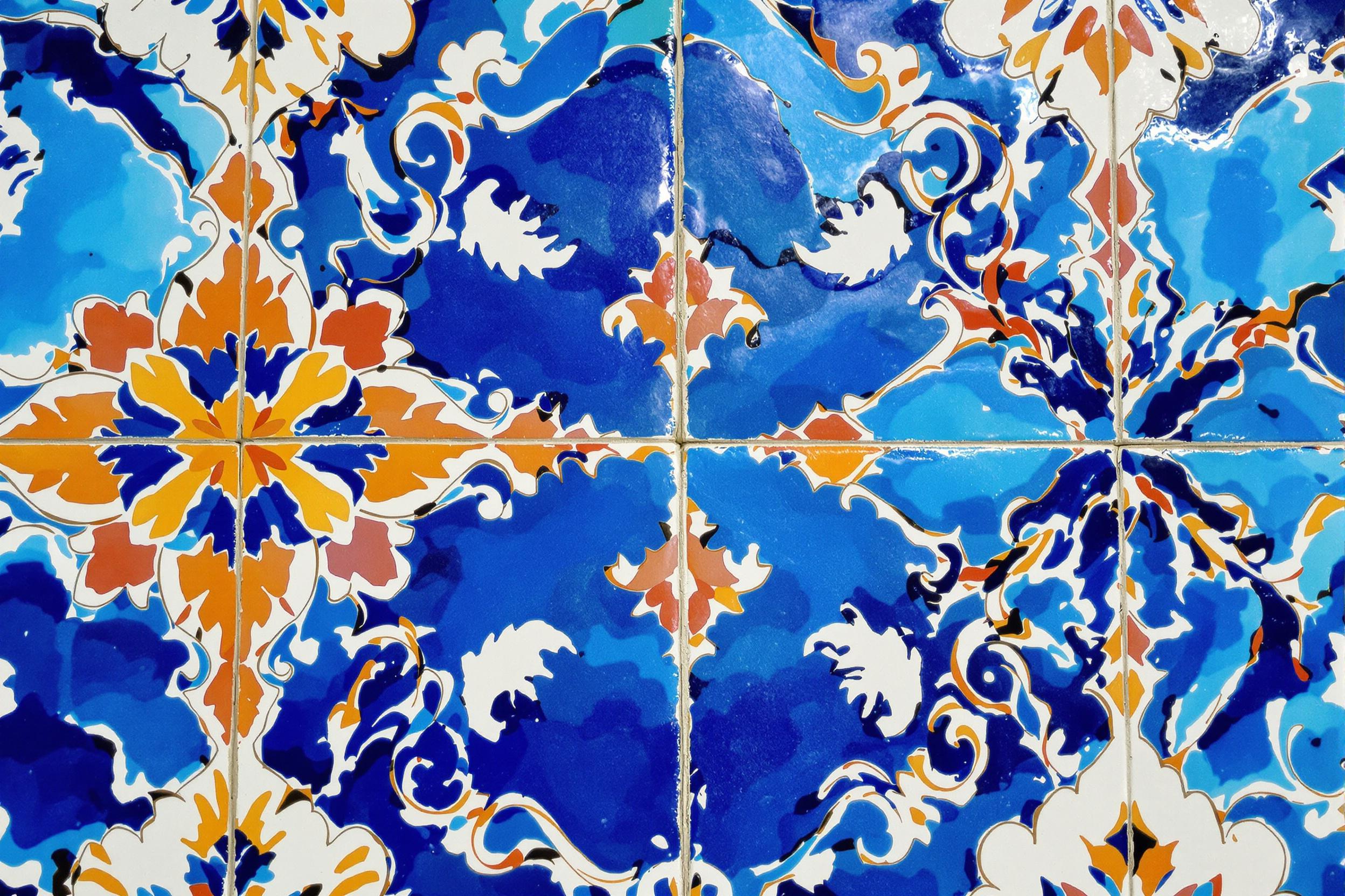 Close-up of vibrant, hand-painted ceramic tiles forming an intricate Mediterranean pattern. Sunlight accentuates the glossy surface, highlighting the interplay of azure blues, warm terracottas, and crisp whites in this mesmerizing texture study.