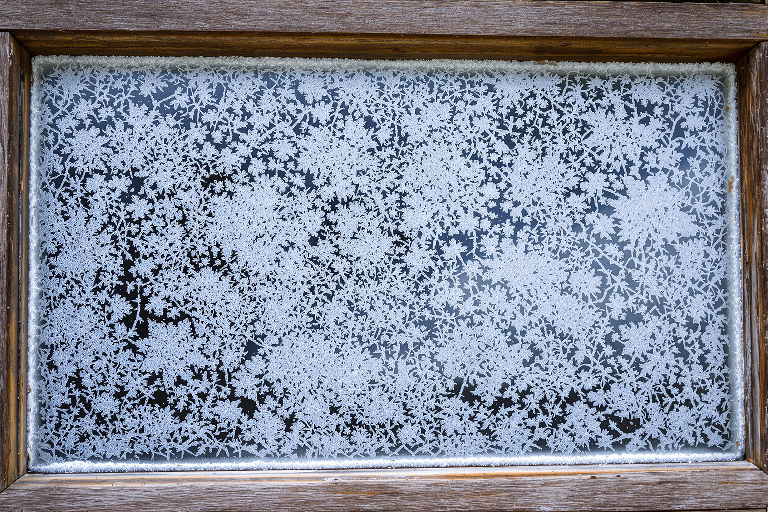 Intricate frost crystals form mesmerizing patterns on an old, weathered window pane. Delicate ice structures create a natural lace-like design, contrasting with the aged wood frame. A perfect blend of nature's artistry and rustic charm.