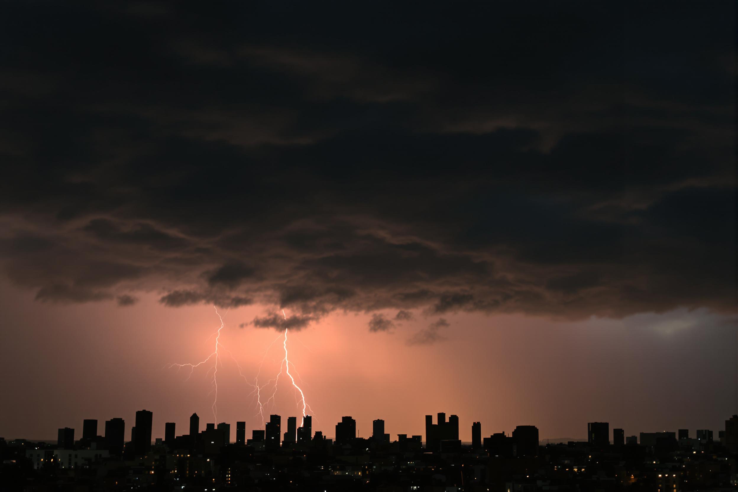 A dramatic thunderstorm looms over a sprawling city skyline at dusk. Dark, swirling clouds fill the sky, illuminated sporadically by flashes of lightning. The silhouette of skyscrapers contrasts sharply against the turbulent backdrop, while the last rays of sunlight create a striking boundary between day and night. Occasional raindrops can be seen falling toward the earth, hinting at the impending downpour.