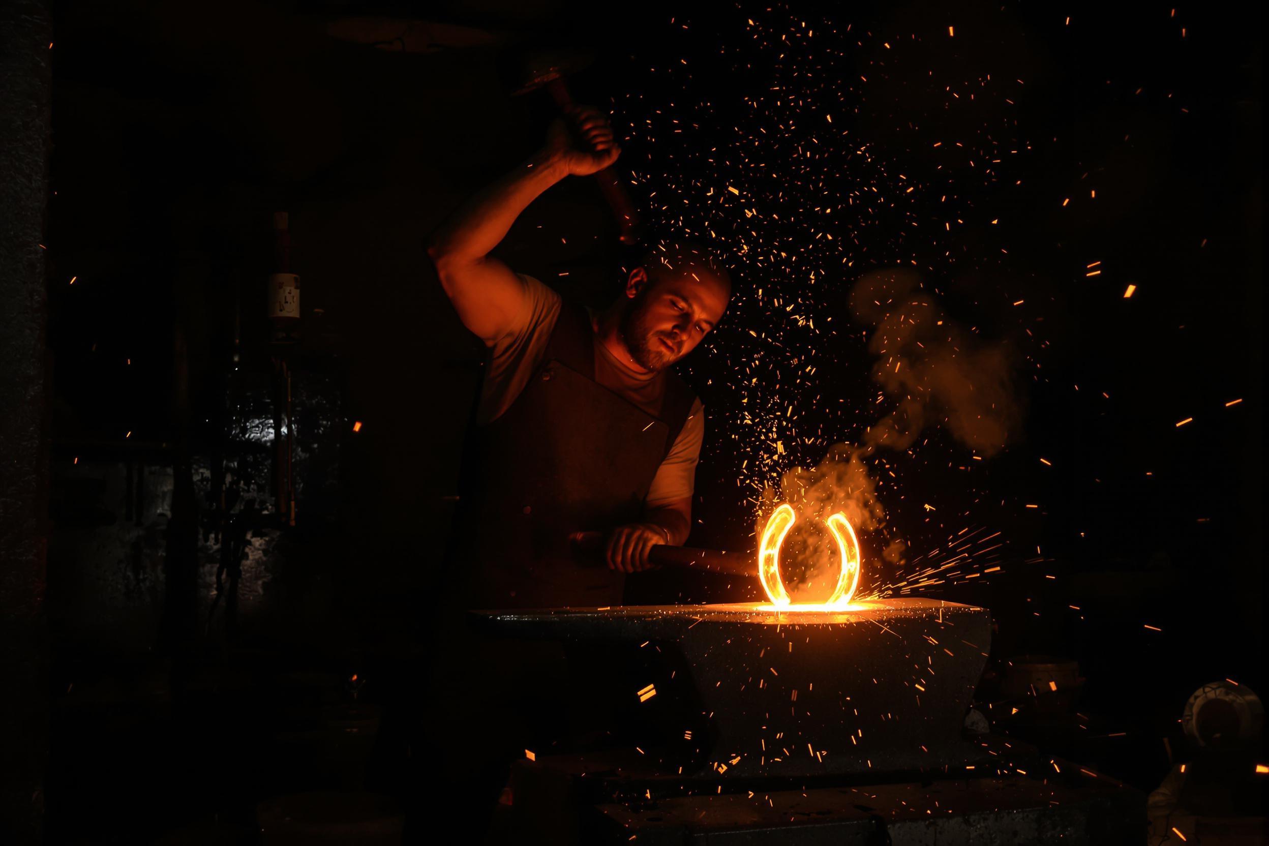 A traditional blacksmith works in a dimly lit forge, illuminated by the warm glow of molten metal. Hammer raised mid-swing, he concentrates on shaping a glowing horseshoe atop an anvil. The room is filled with gritty textures—blackened walls, rustic tools on racks, and rising smoke interspersed with scattered sparks under flickering orange light.