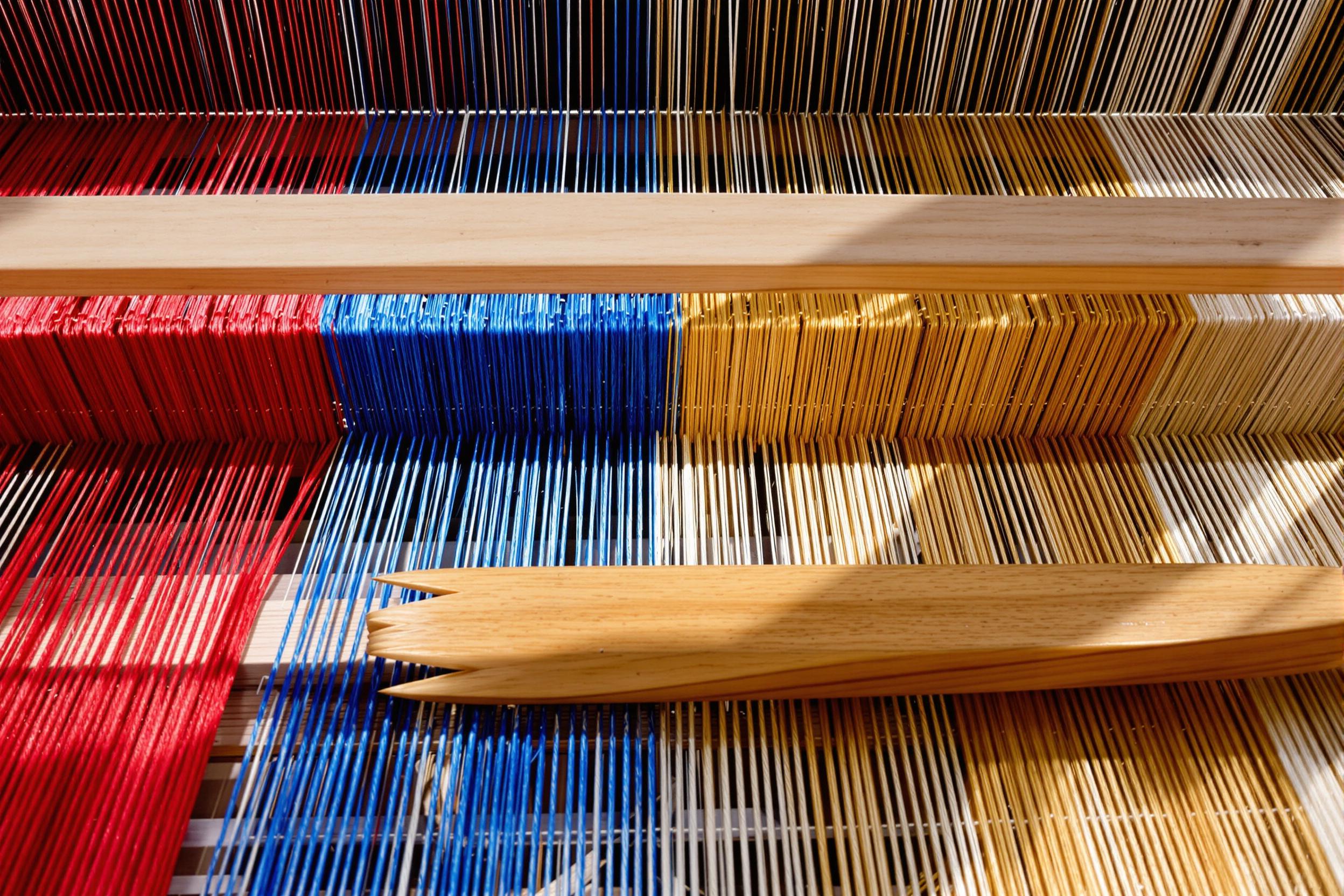 An intricate, close-up view captures a traditional wooden loom mid-operation. Vividly dyed threads—scarlet, azure, and gold—run tautly across the frame, illuminated by slants of natural light from nearby windows. The polished wooden shuttle rests upon textured warp threads while smooth wear marks suggest years of craftsmanship.