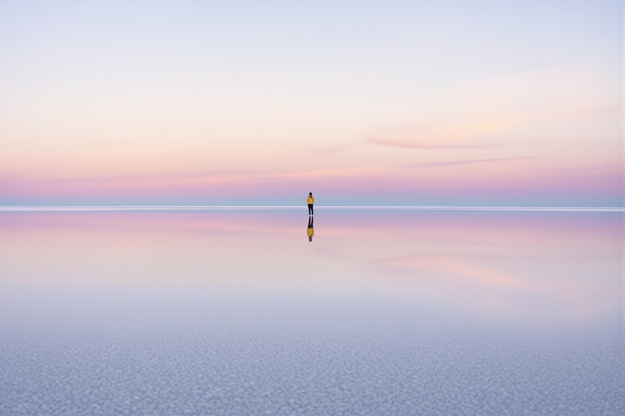 Experience the ethereal beauty of the world's largest salt flat at dawn. A lone traveler stands silhouetted against the pastel sky, perfectly reflected in the thin layer of water covering the crystalline surface. This breathtaking landscape showcases nature's mirror effect, creating an illusion of infinity.