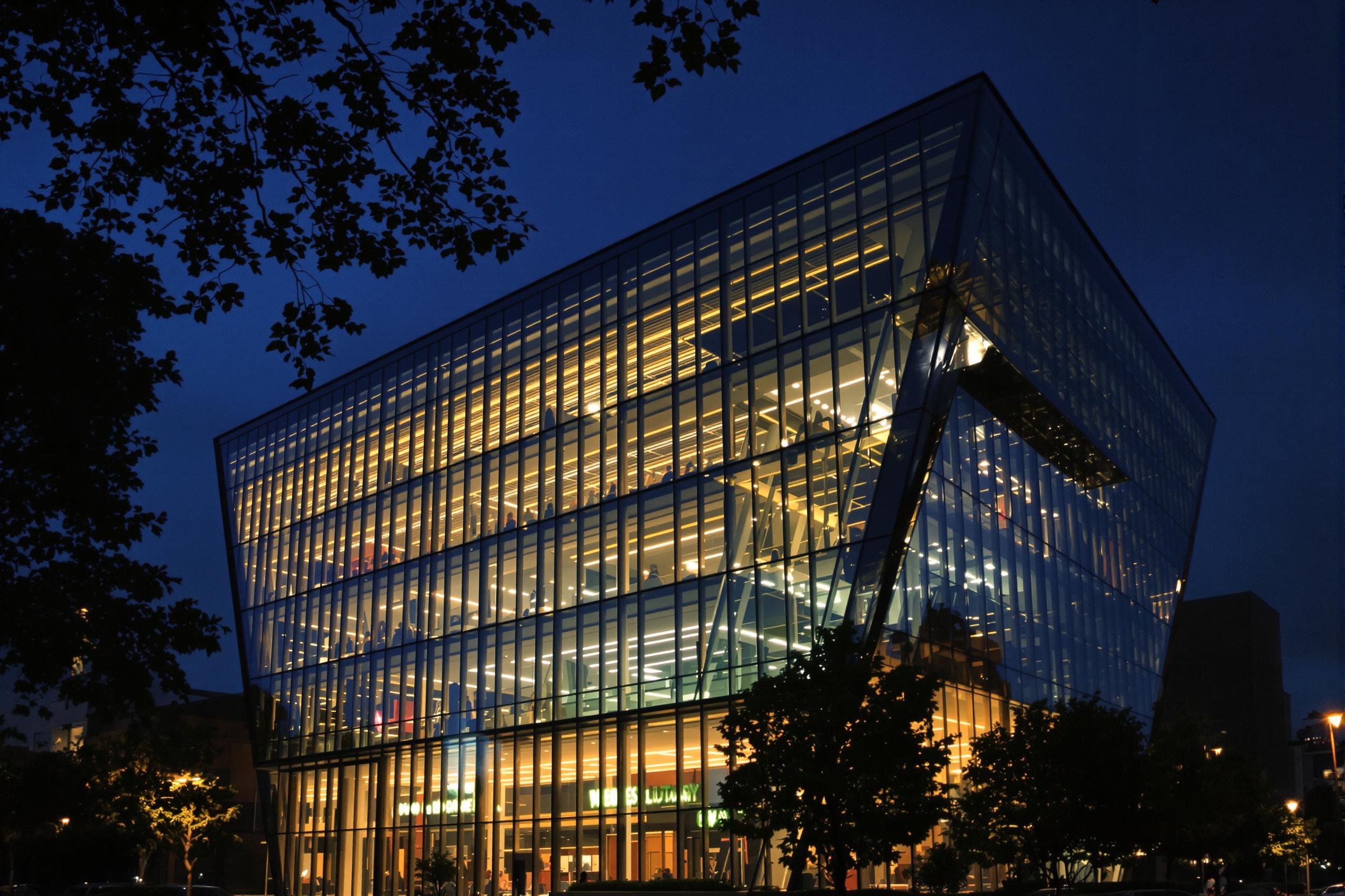 A striking modern library rises in an urban landscape at dusk. Its glass façade reflects the fading day while warm lights spill from within, creating a welcoming glow. The unique angular design features bold lines and shapes that captivate viewers. Surrounding trees add life to the scene, subtly framing the library against a darkening blue sky.