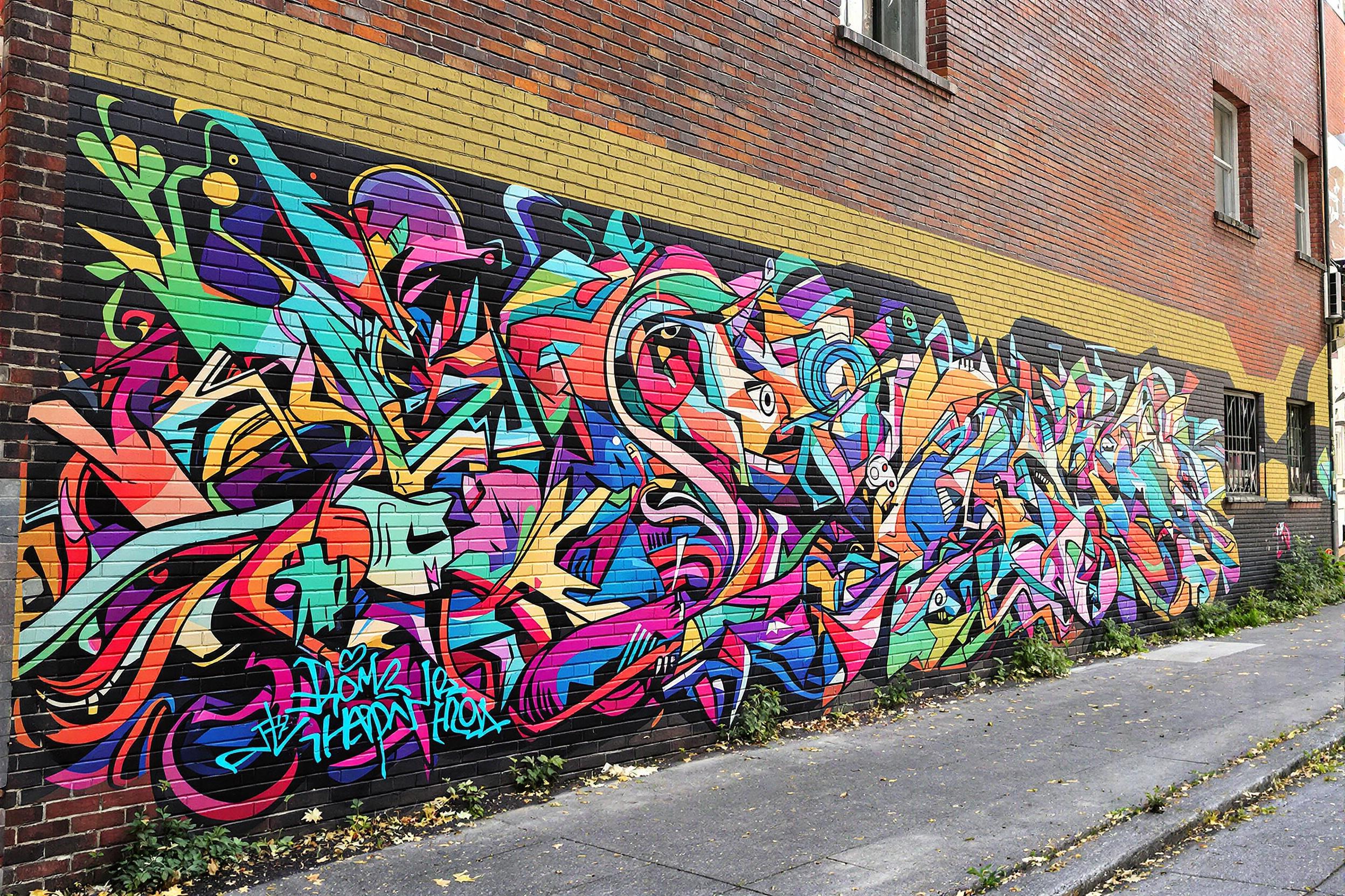 A striking street art mural sprawls across the brick wall of an urban alley, bursting with vivid colors and imaginative designs. The artwork features swirling patterns and abstract forms, illuminated by the soft morning light, highlighting its intricate details. Surrounding the mural, aged brick surfaces add contrast, while scattered greenery peeks through nearby spaces, creating a visually engaging urban landscape.