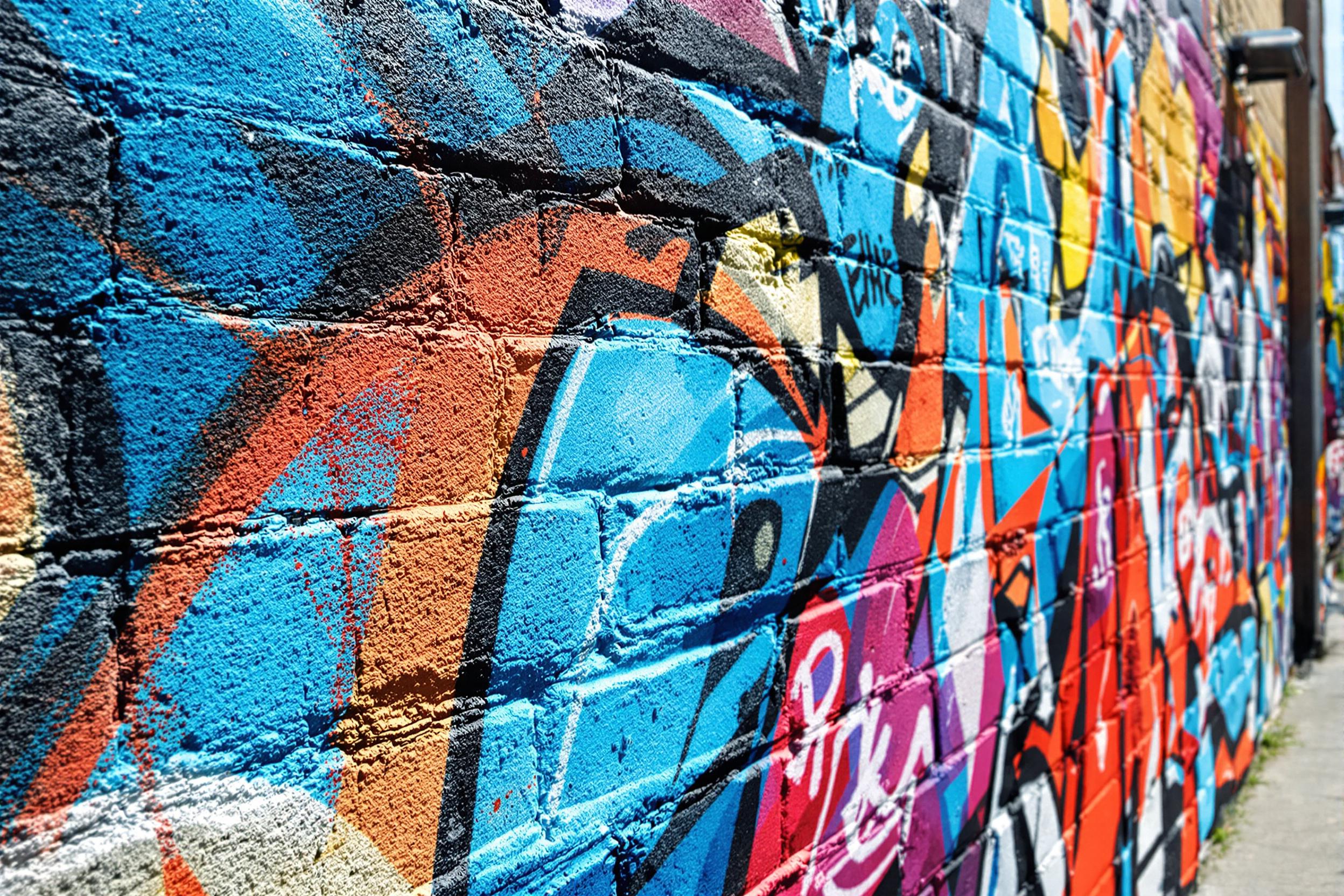 A vivid close-up reveals intricate street art adorning a weathered brick wall. Bright hues of electric blue and fiery red intertwine with energetic patterns and abstract shapes. Textures of spray paint show depth and movement, while sunlight illuminates the vibrant artwork, creating a lively urban atmosphere against a softly blurred gray backdrop.