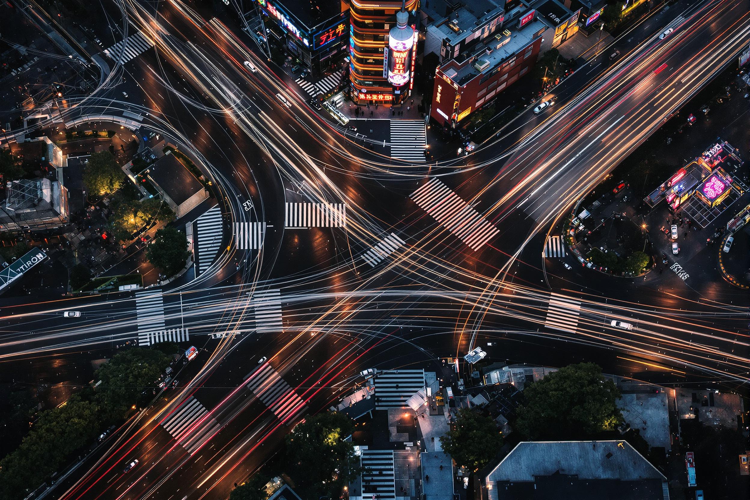 A top-down aerial view captures an intricately networked urban crossroad during peak evening rush hour. Streams of headlights create dynamic streaks against dark asphalt, highlighting a symphony of movement. Neon signage from surrounding buildings reflects off rain-slick pavements, while well-lit pedestrian crossings weave human-scale order into the scene.