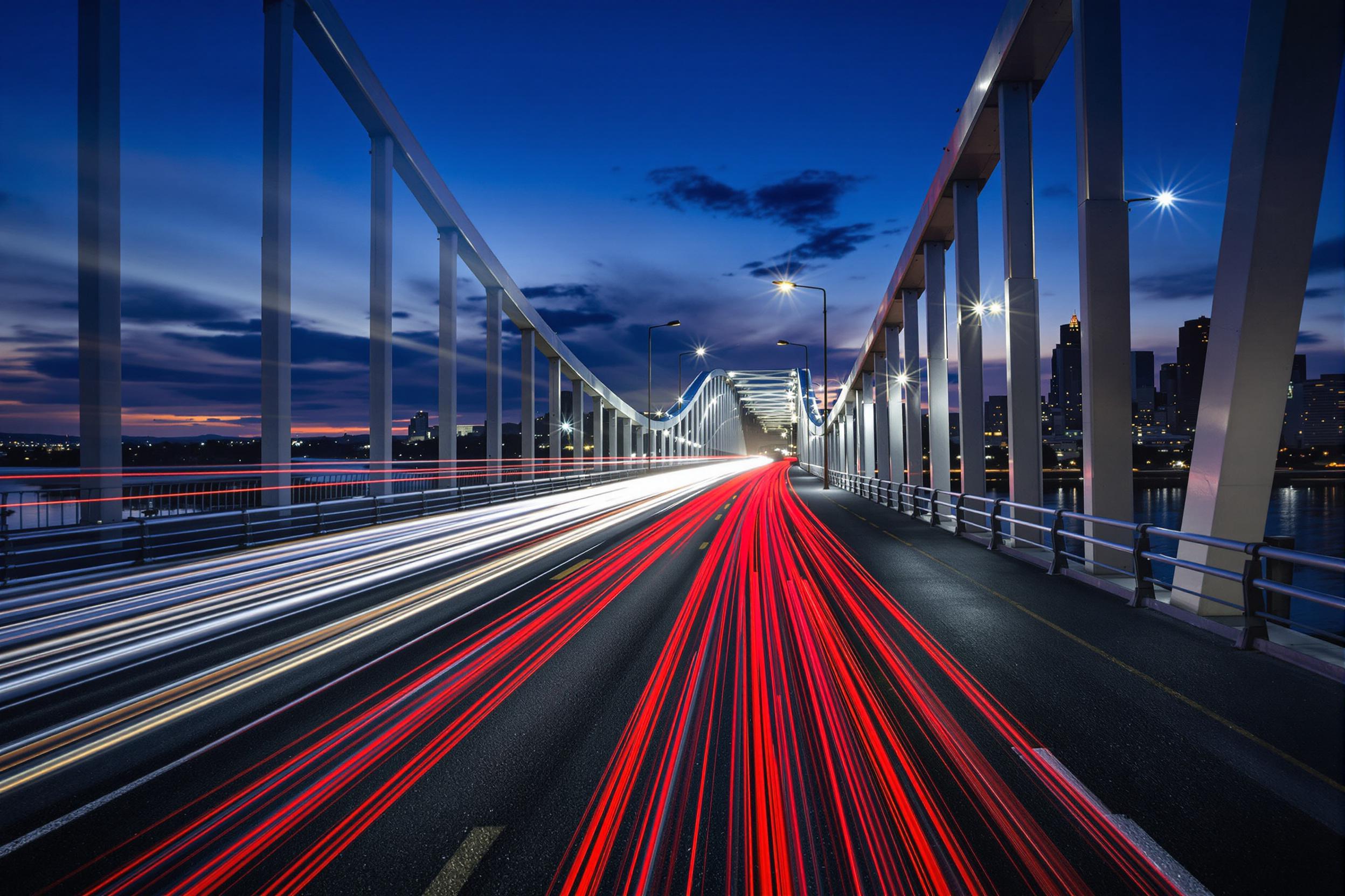 An urban bridge comes alive at twilight as vehicle light trails paint the scene. Red and white streaks of light from cars weave above dark asphalt, bordered by sleek railings. The glow of the city skyline and fading daylight adds depth, creating a vivid display of motion and color.