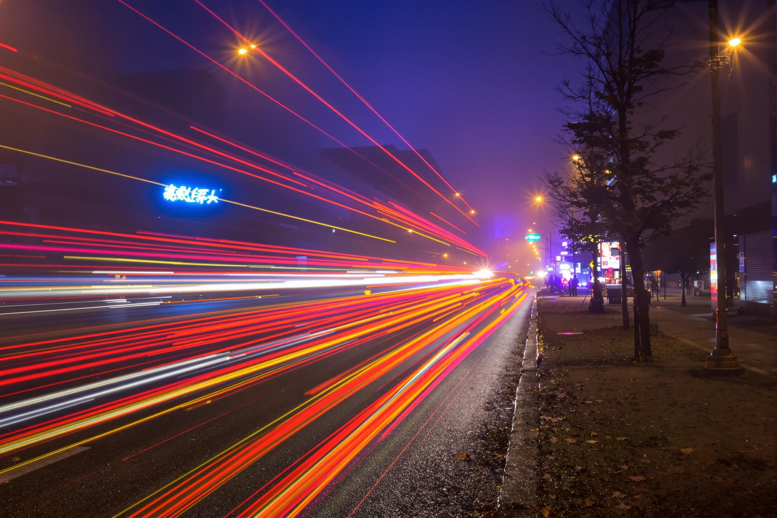 Dynamic light trails from vehicles stream smoothly across a foggy urban street at night. Long exposure captures vivid streaks of red, gold, and white that weave through soft blue and purple mist. Hints of neon signs and urban architecture peek faintly in the background, adding depth and vibrancy against the hazy nighttime atmosphere.