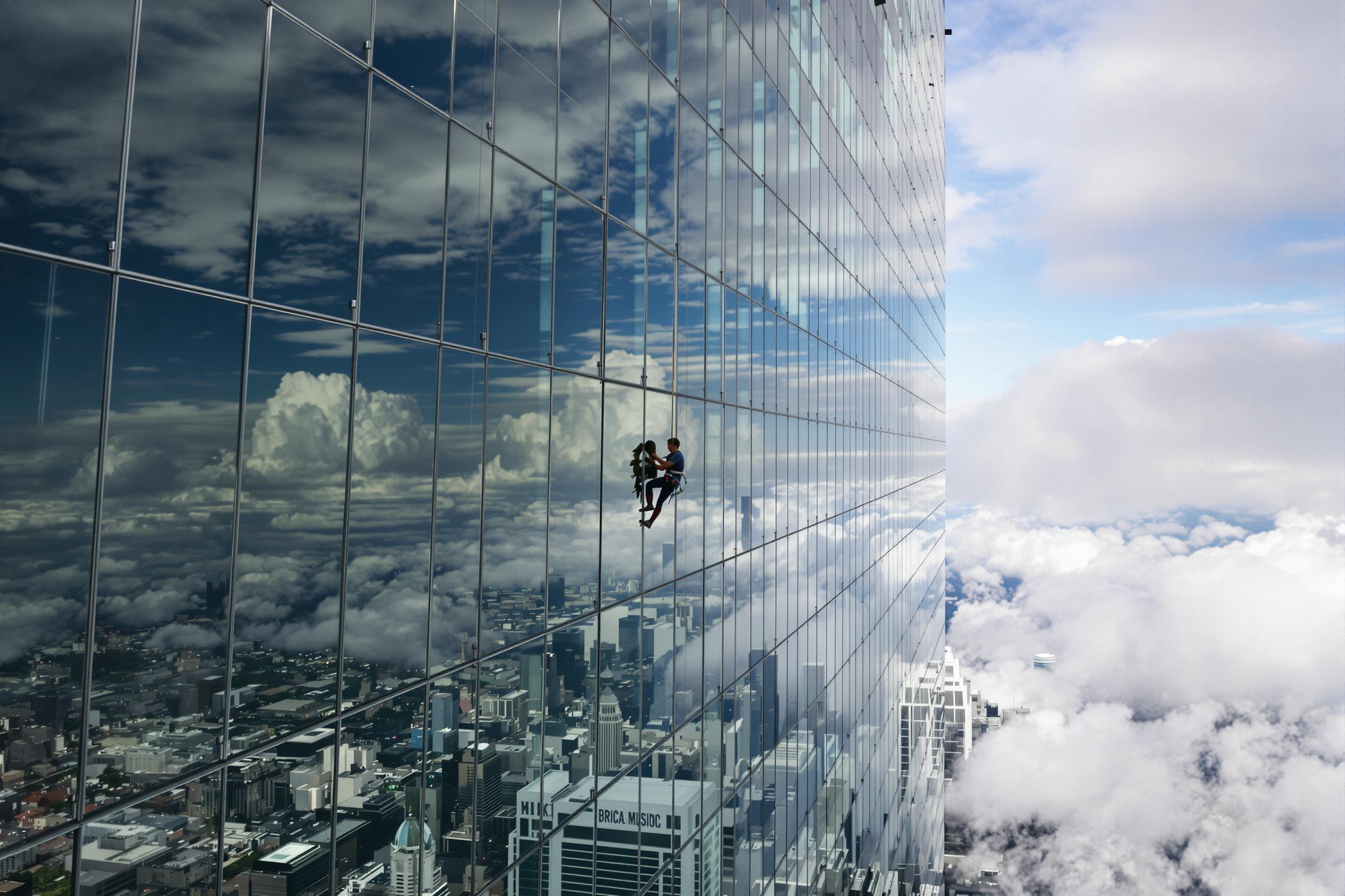 Against the backdrop of a sprawling urban skyline, a lone climber hangs from the reflective surface of a glass skyscraper. Cloud patterns reflect on the building’s facade, blending with its geometric structure. Viewed from a worm's-eye angle, the perspective amplifies the dizzying verticals, while soft lighting casts gentle highlights enhancing the sense of height.