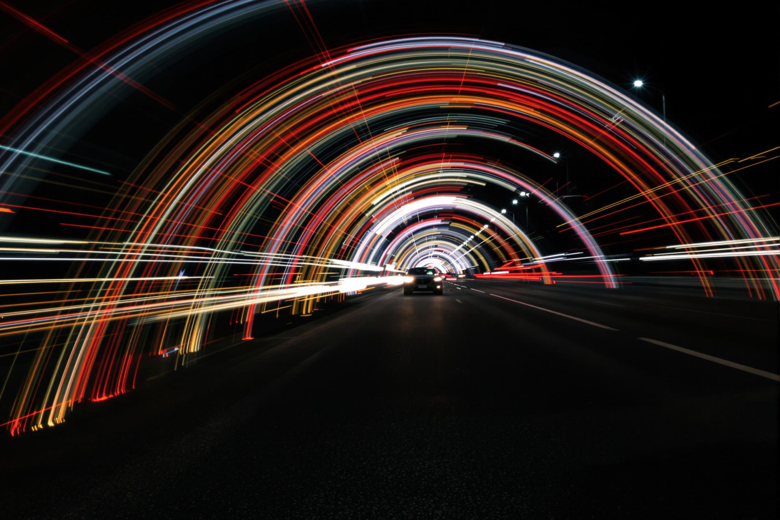 Dynamic arcs of colored light trail through a bustling urban street scene, captured via long-exposure photography. Blurred traffic creates radiant streaks of red, yellow, and white against dark asphalt. Distant city lights glow, while faint silhouettes of passing cars suggest movement.