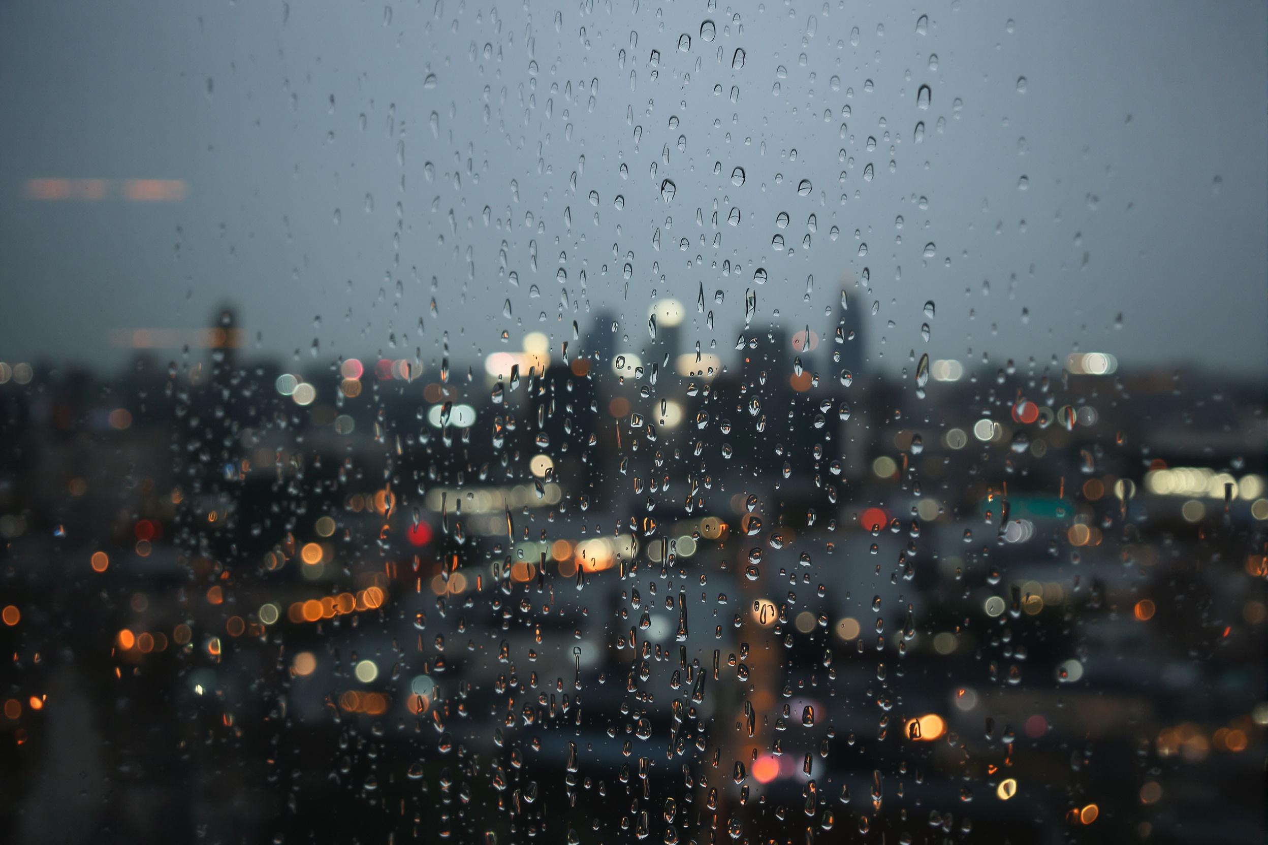 Raindrops streak down a glass window as a city skyline glimmers softly under the fading light of dusk. Blurred buildings and shimmering lights provide a muted backdrop, while vibrant reflections dance along the glass, creating an intimate yet dynamic composition.