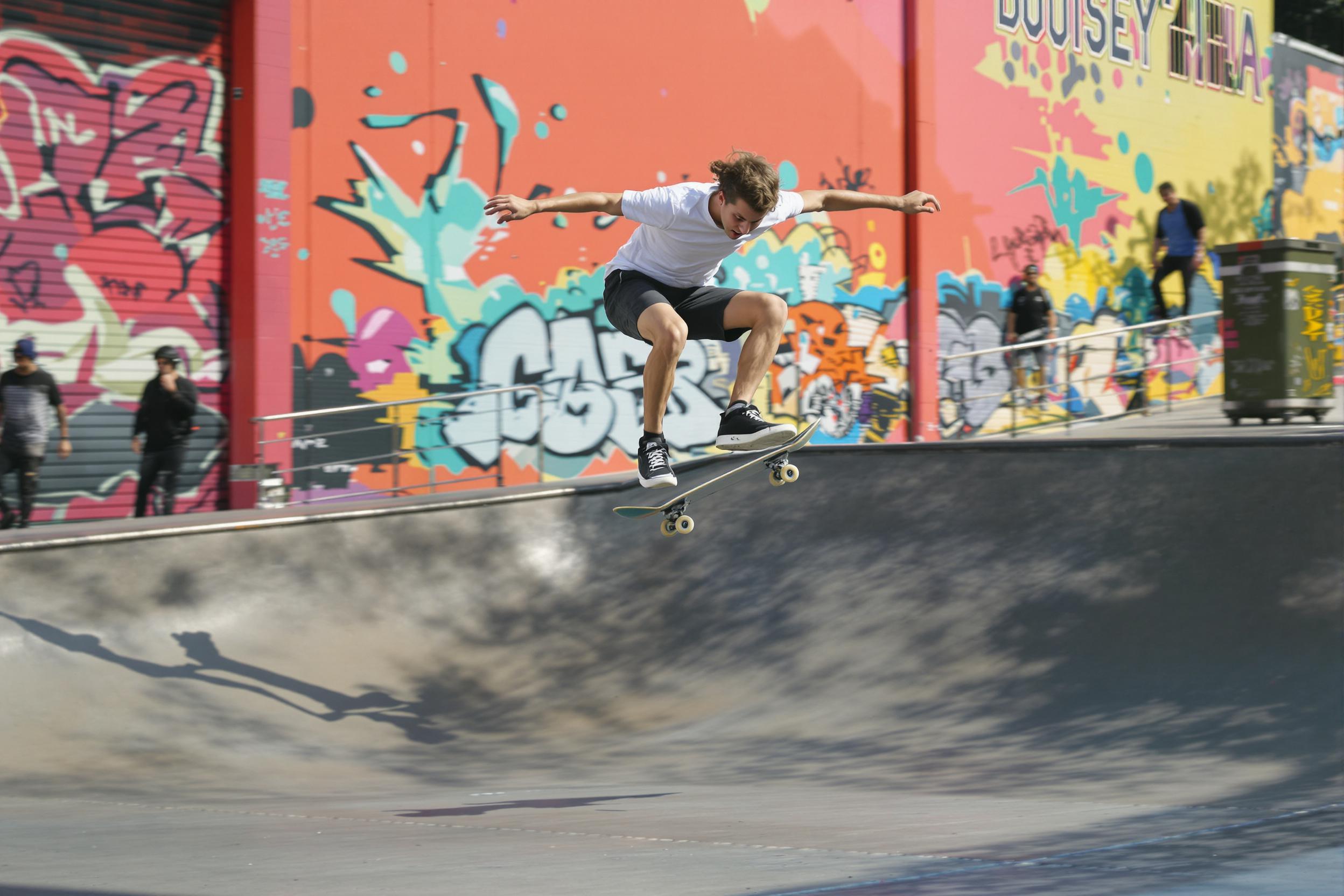 A teenager captured mid-air during a dynamic skateboarding trick in an urban skatepark setting. Bright afternoon light casts long shadows on smooth asphalt while vibrant, graffiti-splashed walls in the background frame the action. Motion-blurred silhouettes of other skaters emphasize both speed and energy in the scene.