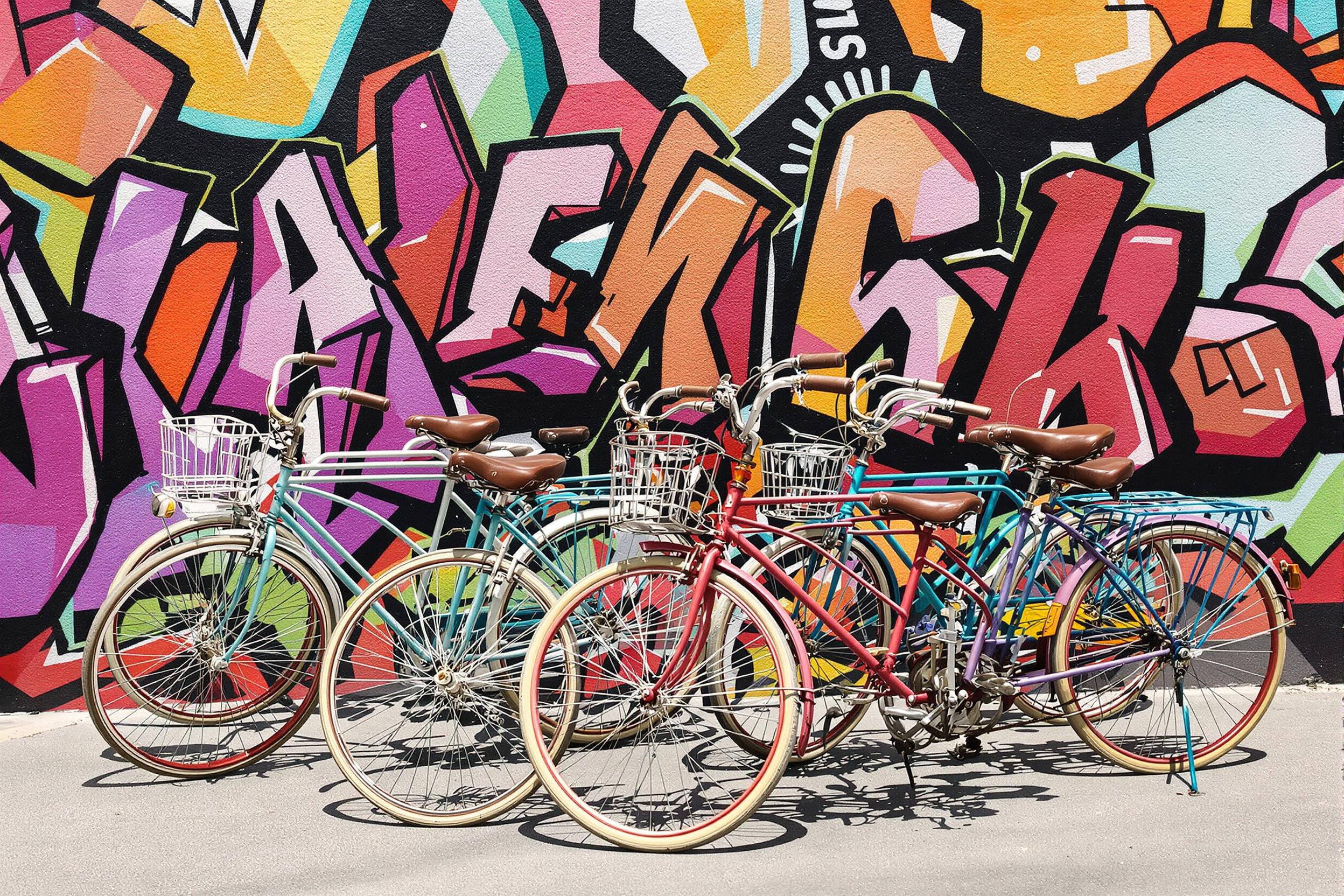 A cluster of vintage bicycles stands proudly against a bold, graffiti-covered wall in an urban setting. The bikes, each unique in color and style, showcase intricate details such as leather saddles and spoked wheels. Sunlight illuminates their glossy surfaces, while the vibrant mural provides a lively backdrop, enhancing the charm and nostalgia of this scene.