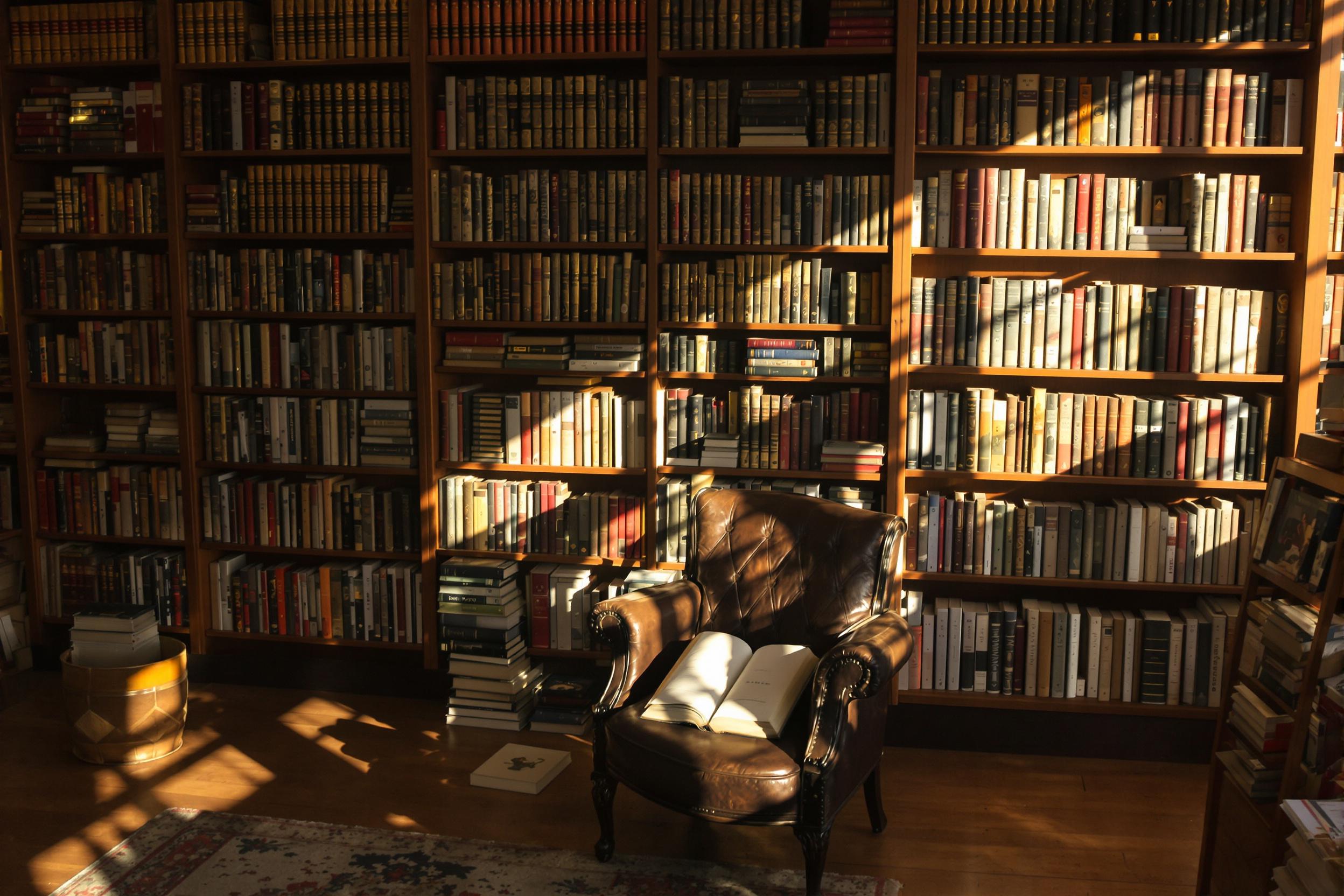 Warm evening sunlight streams diagonally into a vintage bookstore, illuminating rows of aged hardcovers atop wooden shelves. Near the center, a worn leather armchair sits beneath stacked books, an open tome resting on its seat. Shadows create dramatic patterns across the rich oak flooring, accentuating the store’s rustic charm.