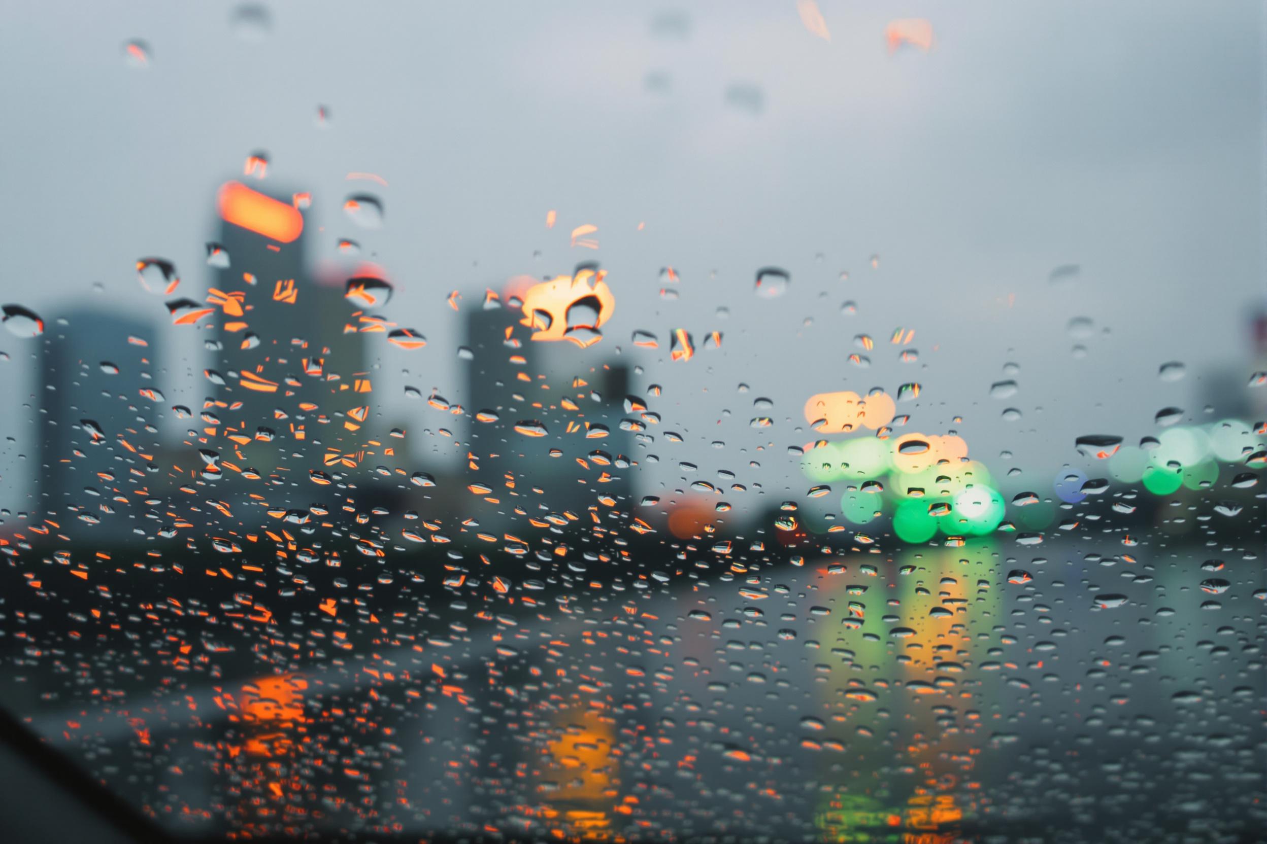 A close-up of large water droplets streaking down a slanted car windshield as urban skyscrapers loom faintly in the blurred background. Dim storm clouds set the tone, while orange and green neon reflections bounce off the rain-slicked surface, imbuing the image with a moody interplay of natural and artificial elements.