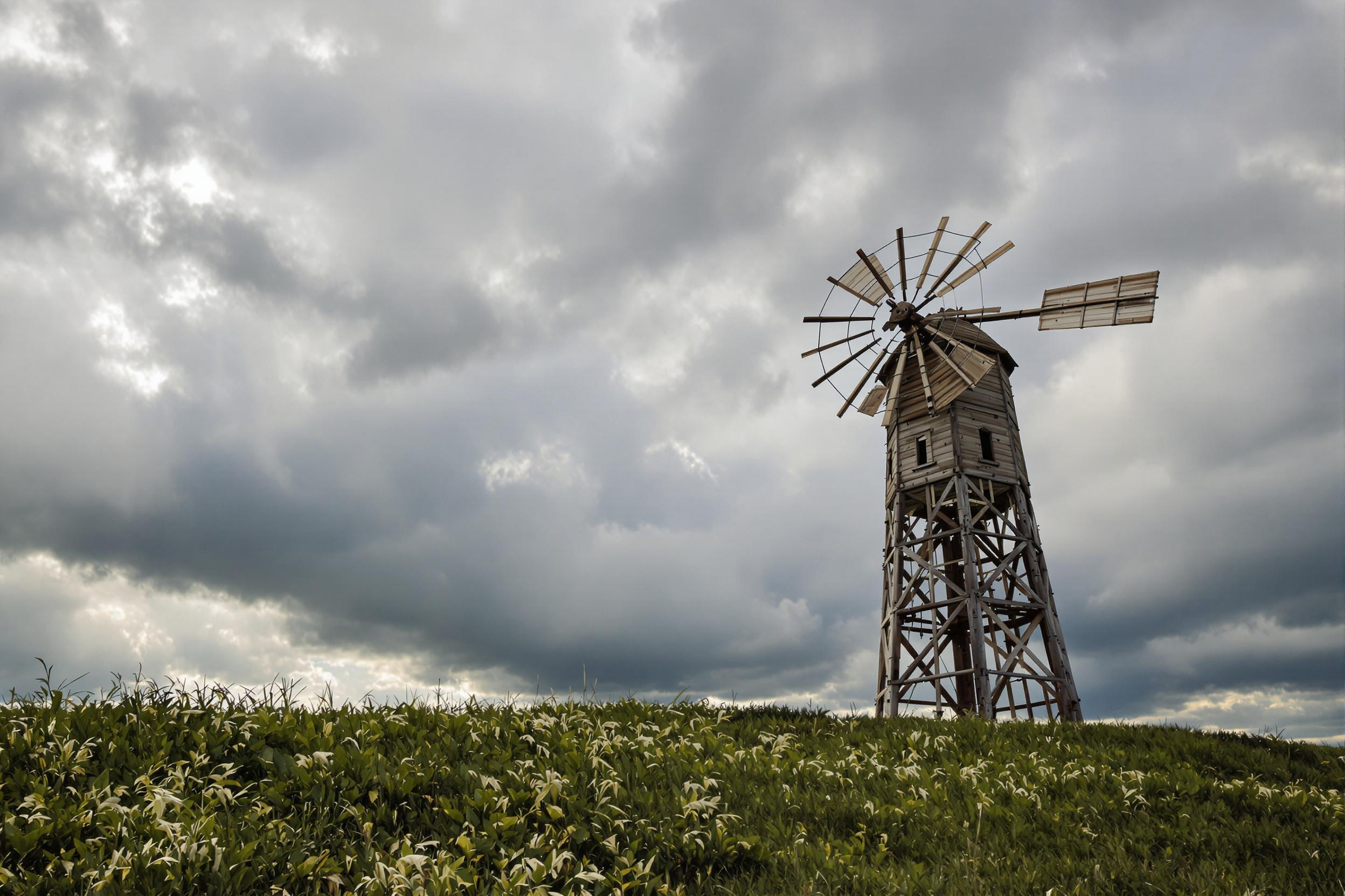 A towering wooden windmill stands alone on a grassy countryside hilltop, dwarfed by towering storm clouds sweeping across the sky. Sunlight faintly breaks through the clouds, casting soft highlights on the aged wooden blades. The ominous weather contrasts with the warmth of the green grass below.