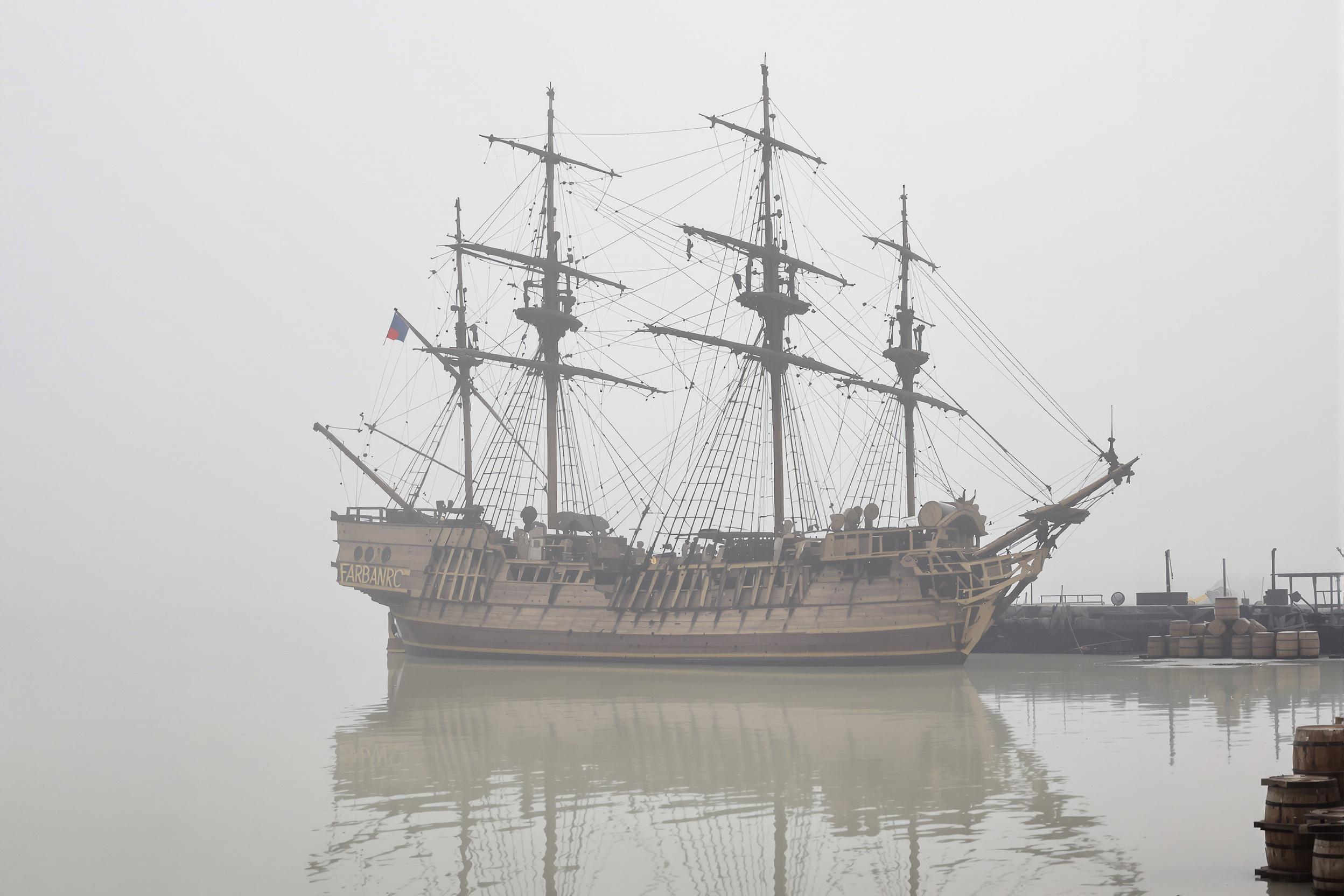 An intricate wooden ship rests quietly in a fog-laden harbor. Towering masts rise against a soft gray sky, draped in weathered sails and complex rigging. The ship reflects faintly in calm, pale waters, while scattered wooden barrels and shipping crates near the dock complete the scene's historical ambiance.